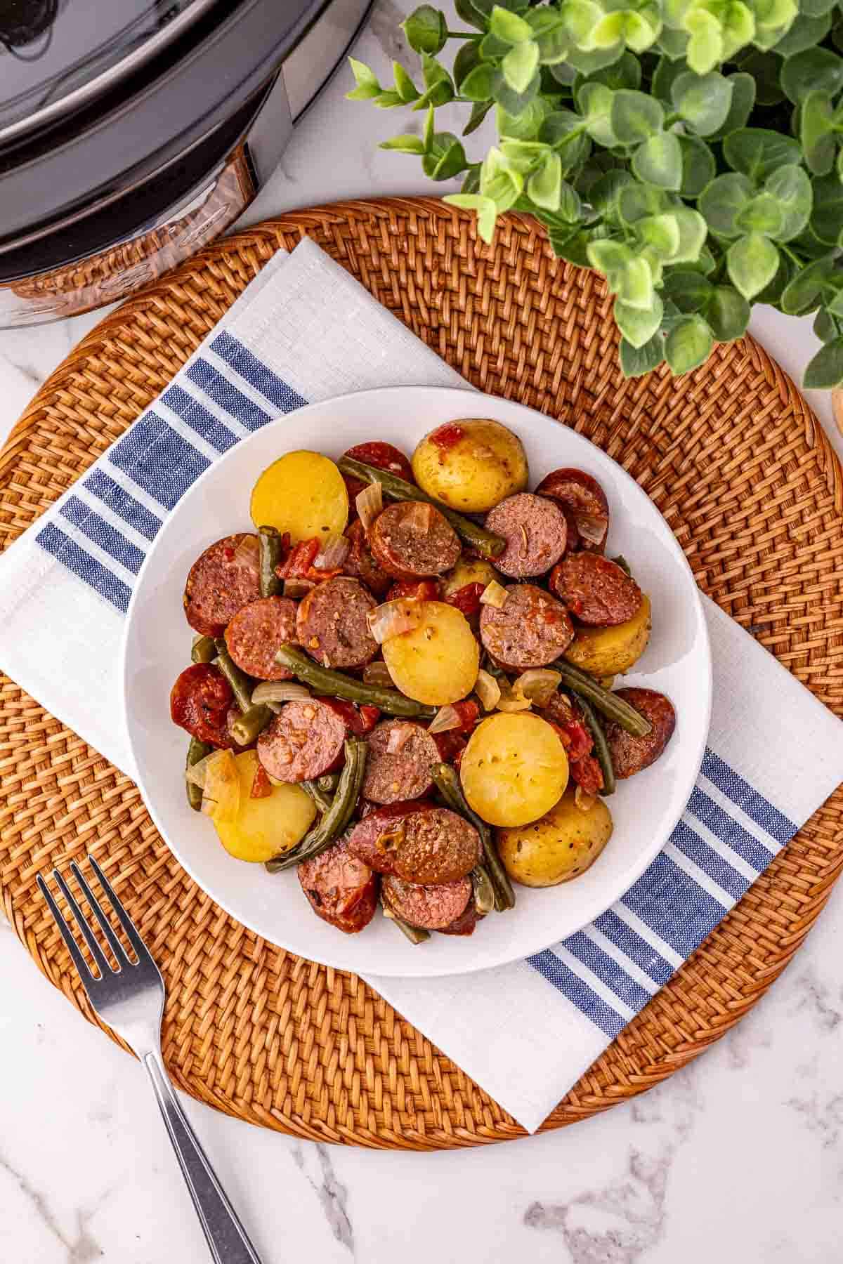 A plate of crockpot sausage, potatoes, and green beans sits on a woven placemat beside a fork and a potted plant.