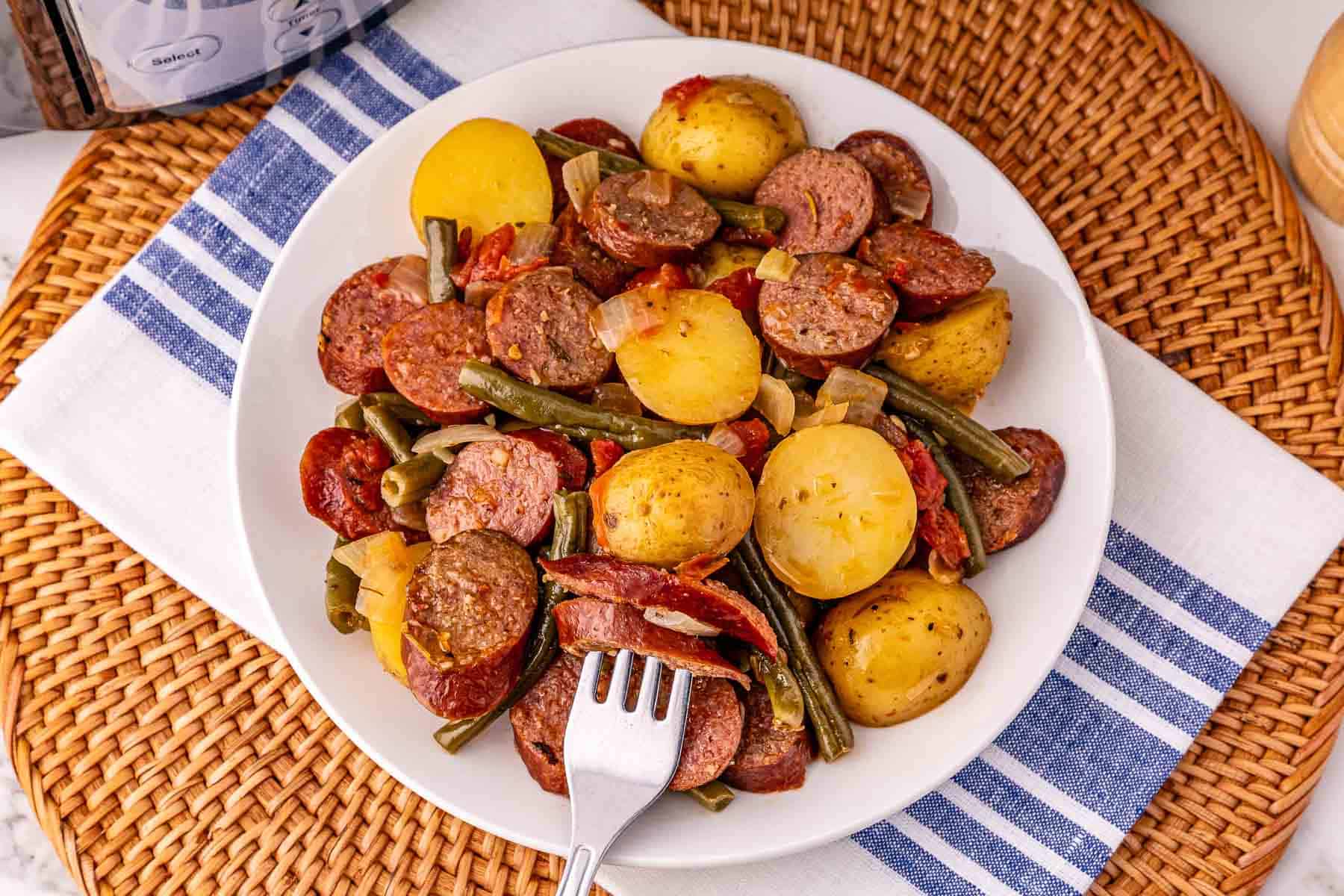 A plate of crockpot sausage and potatoes with green beans and onions sits on a striped napkin, a fork resting in the food.