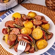 A plate of sausage and potatoes, green beans, and tomatoes sits beside a crockpot of sausage and potatoes and a potted plant.