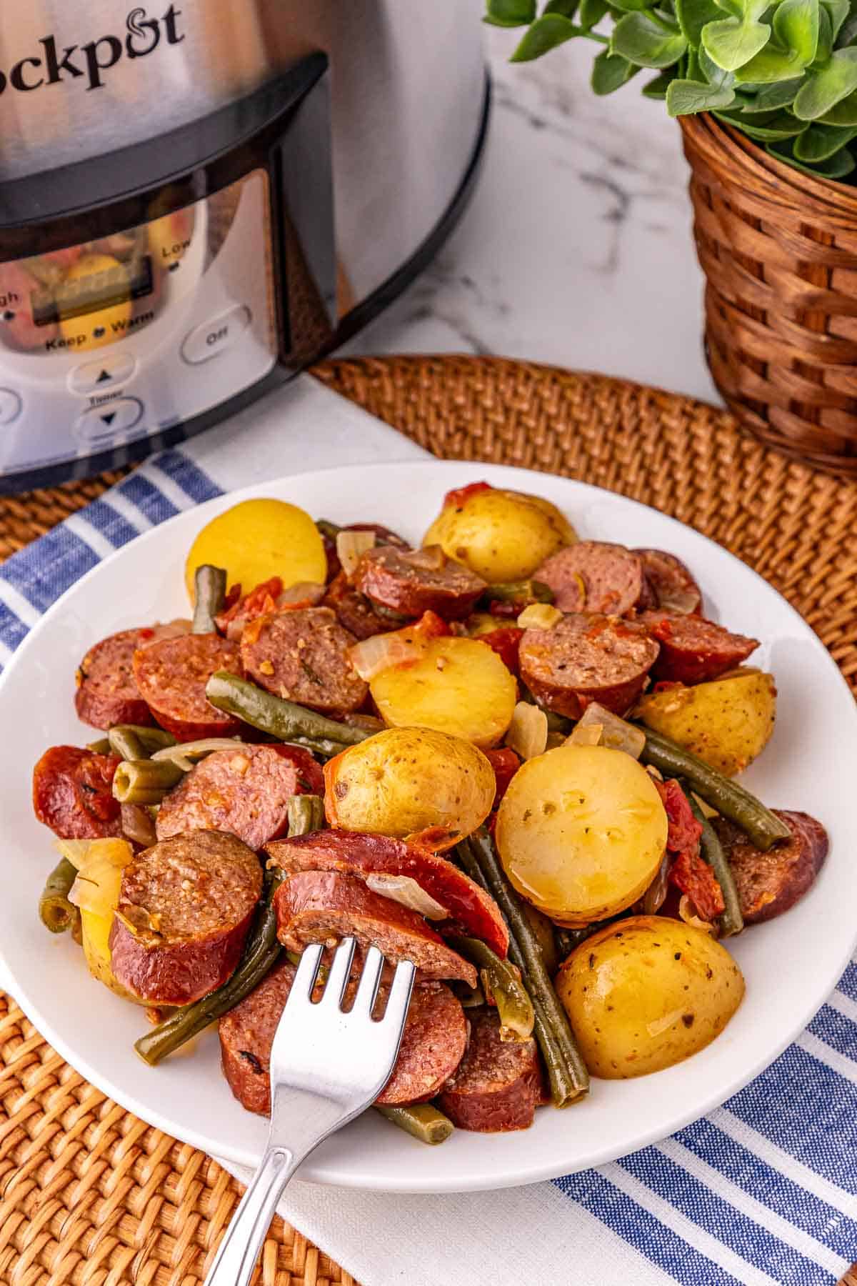 A plate of sausage and potatoes, green beans, and tomatoes sits beside a crockpot of sausage and potatoes and a potted plant.