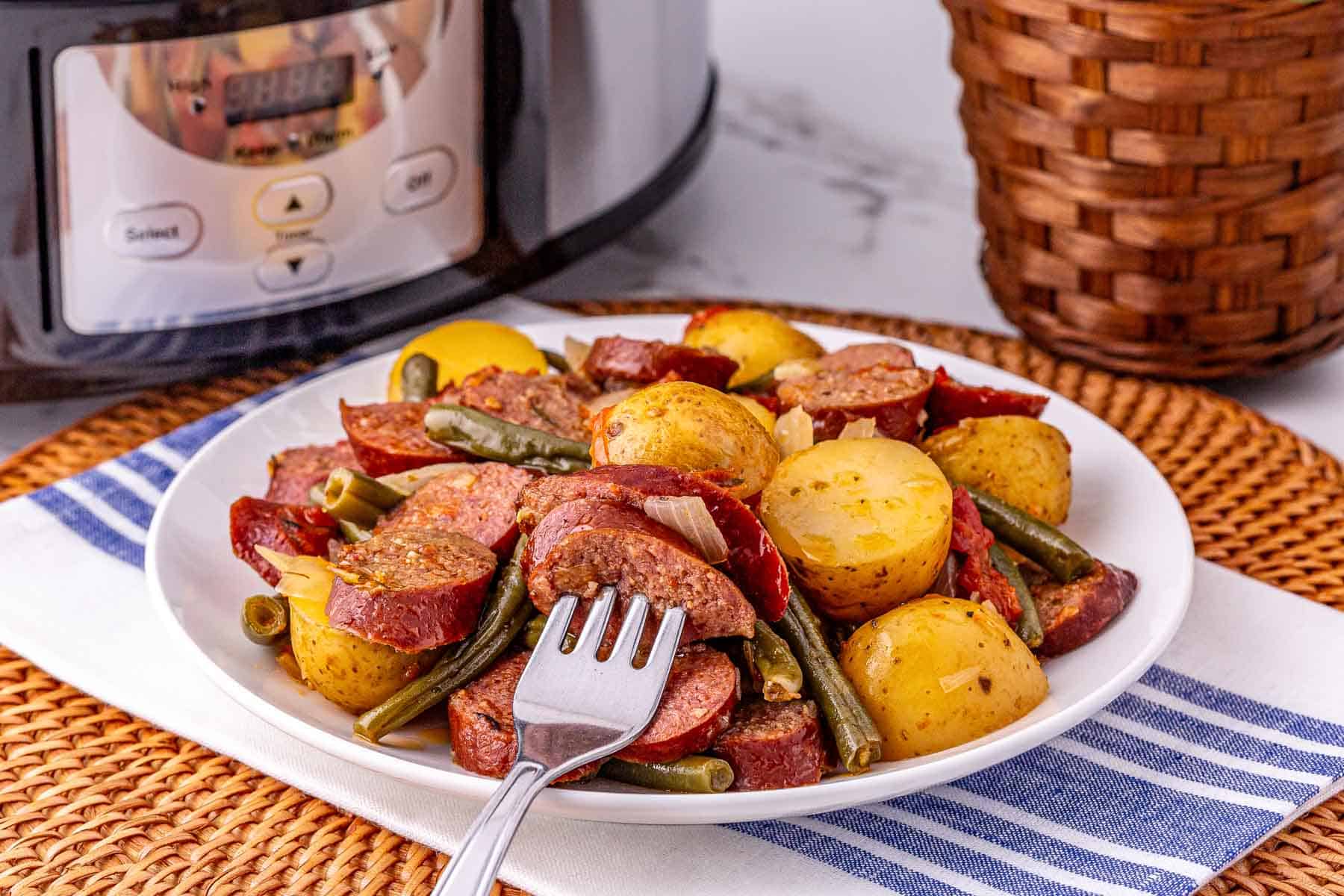 A plate of crockpot sausage and potatoes, green beans, and onions sits in front of a slow cooker on a woven placemat.