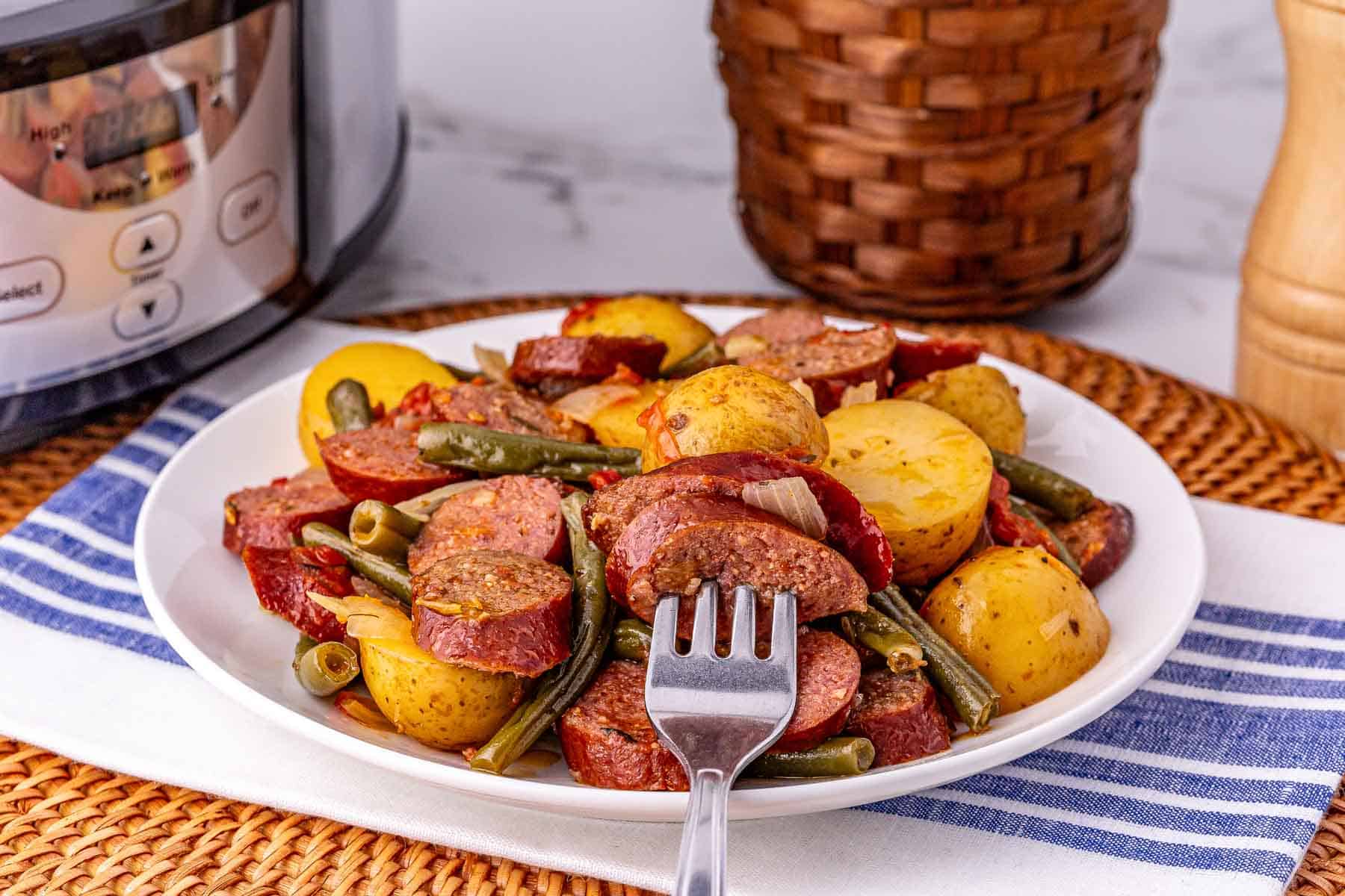 A plate of crockpot sausage, potatoes, and green beans with a fork, set beside a slow cooker and woven basket.
