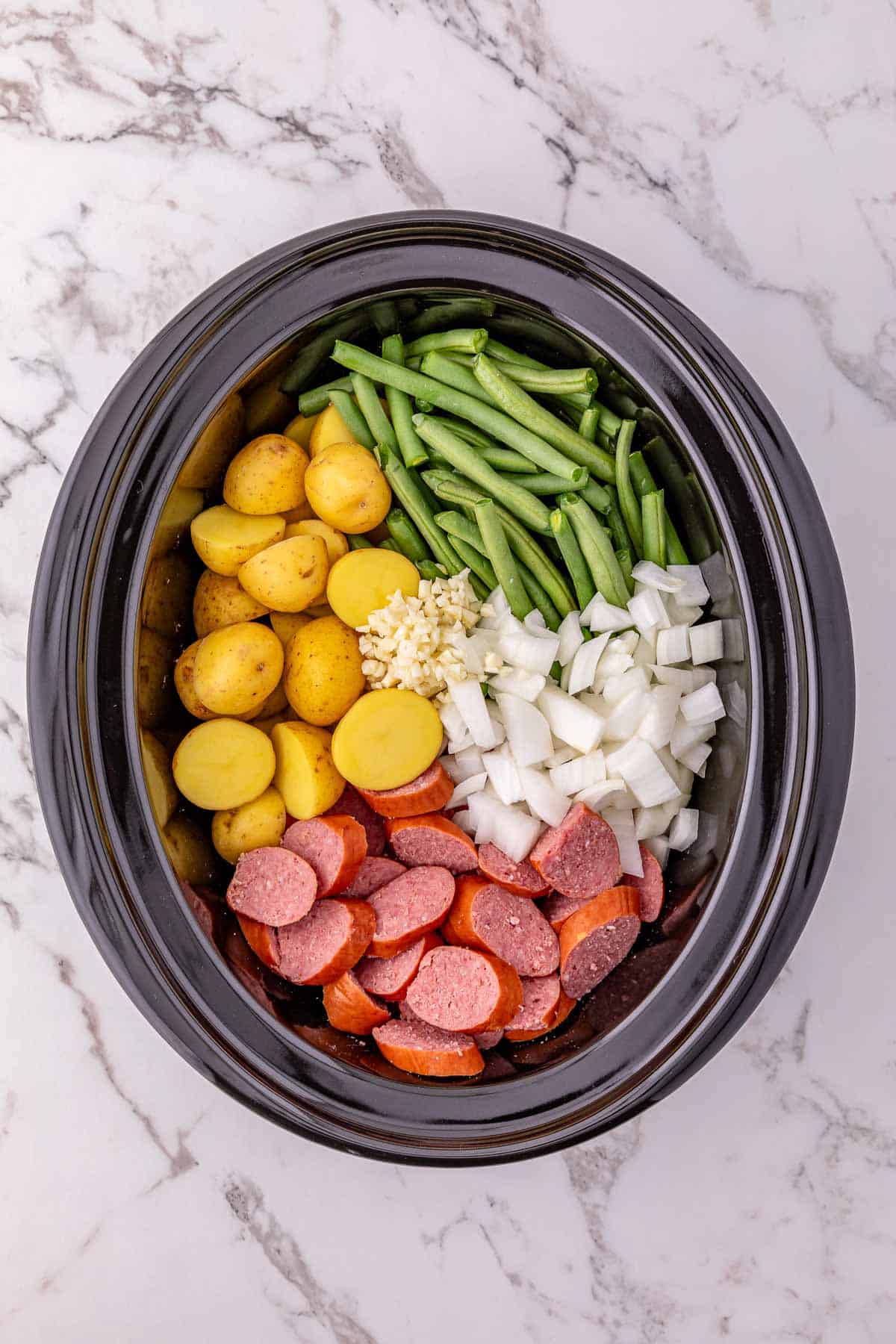 Slow cooker filled with crockpot potatoes, green beans, chopped onions, sausage, and minced garlic for a hearty meal.