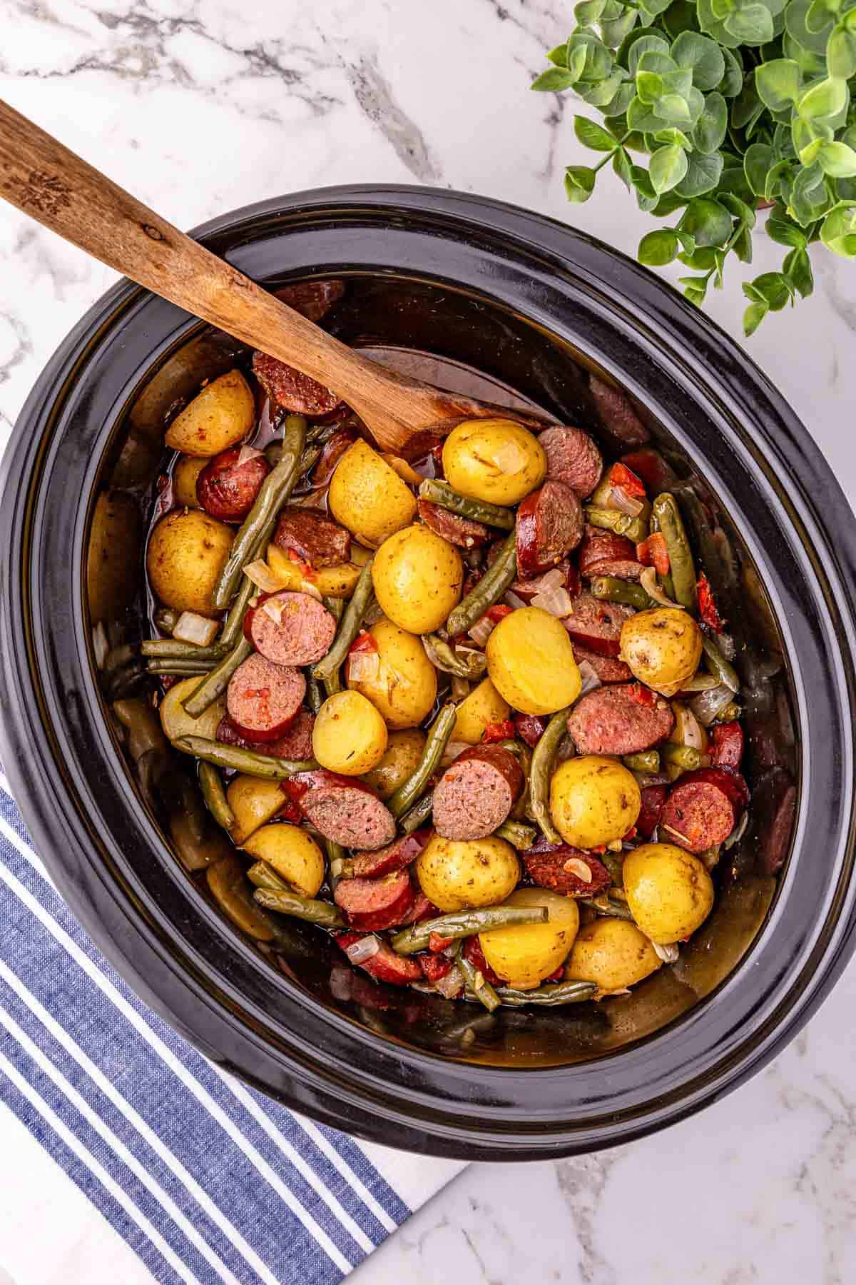 Sliced Crockpot sausage, potatoes, and green beans in a slow cooker with a wooden spoon, on a marble counter.