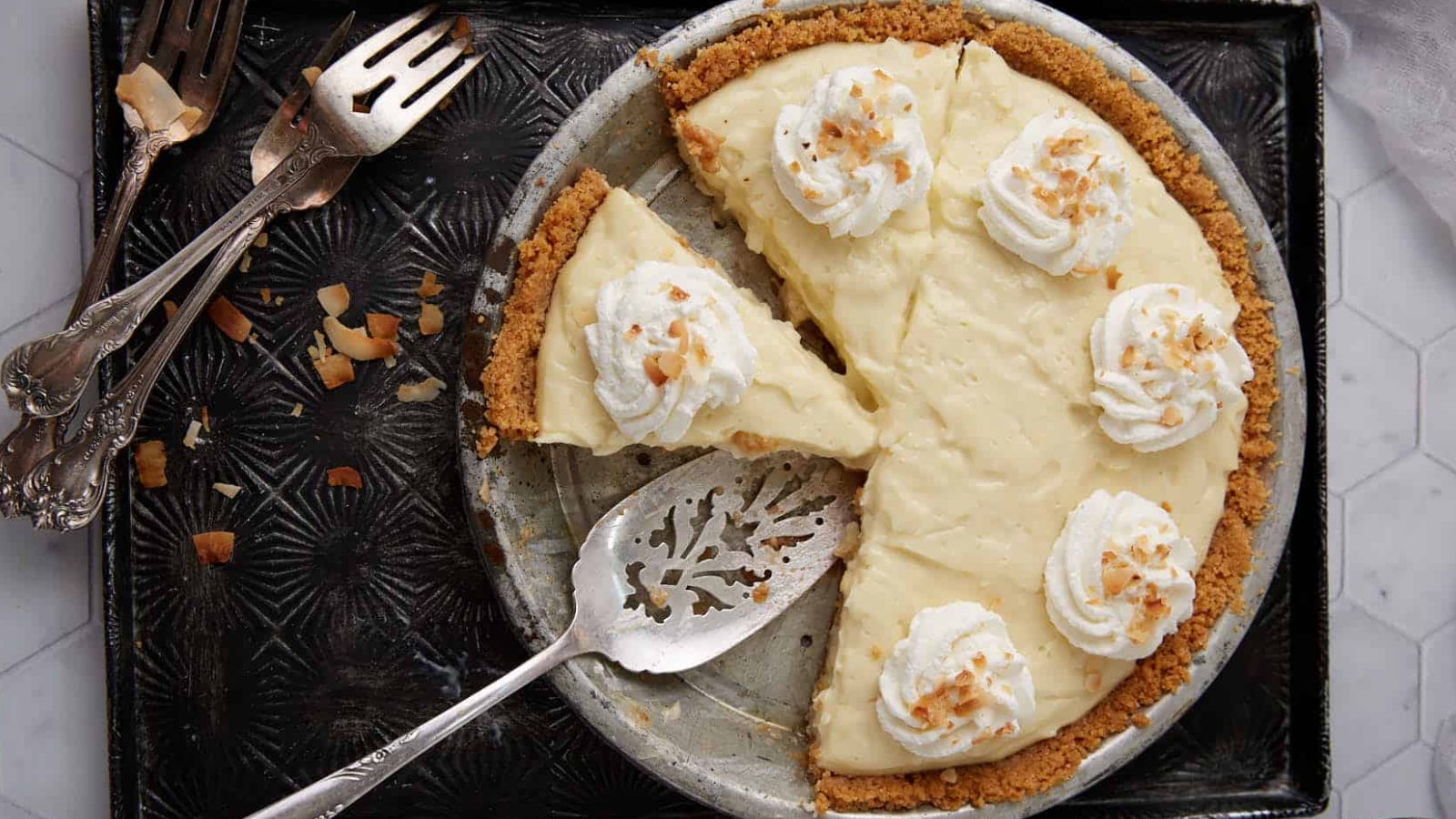 A cream pie with whipped topping and a slice missing, on a tray with serving utensils beside it&mdash;perfect for sharing classic vintage recipes at your next gathering.