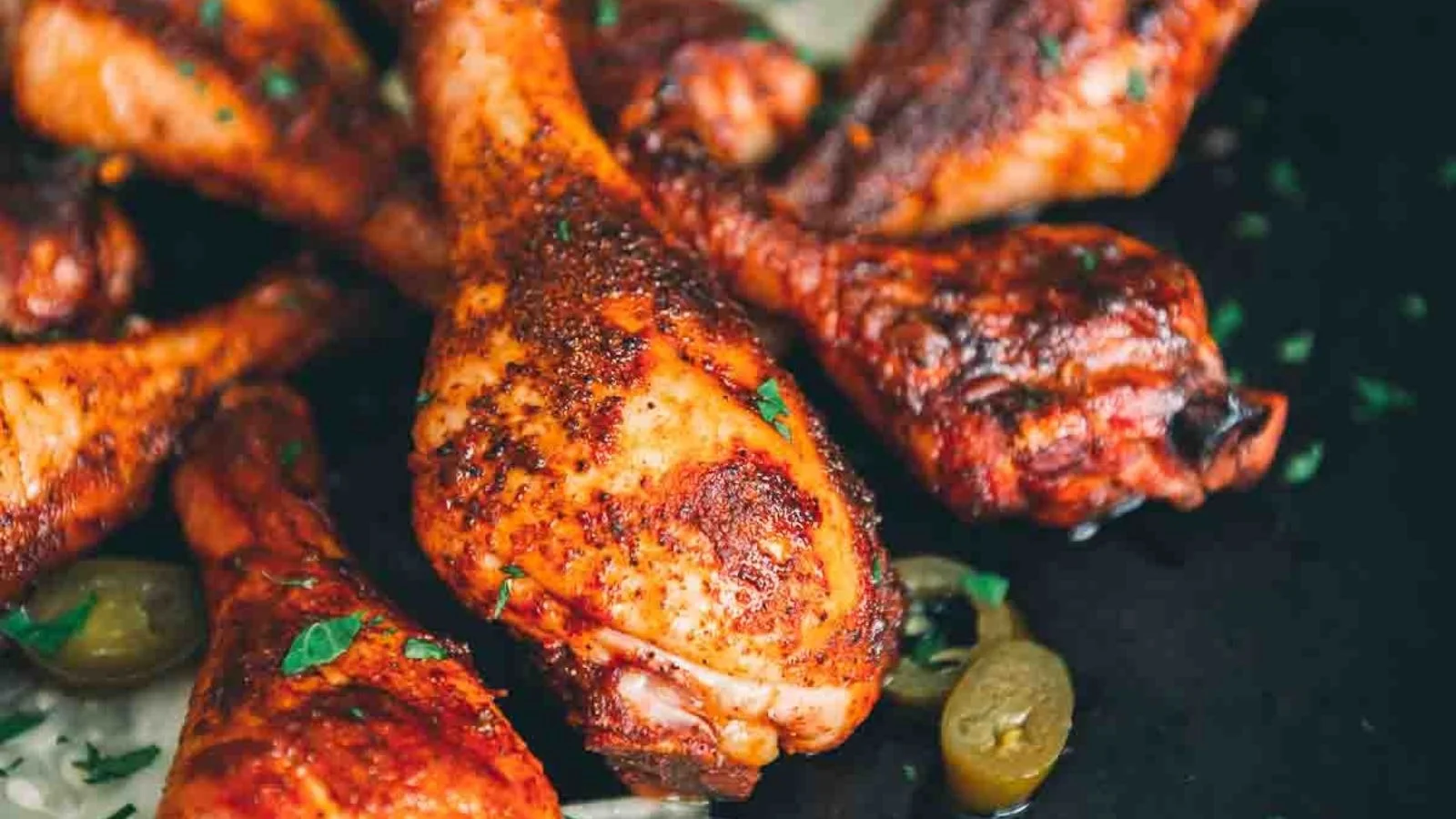 Close-up of seasoned, baked chicken drumsticks garnished with herbs, with sliced jalapeños nearby.