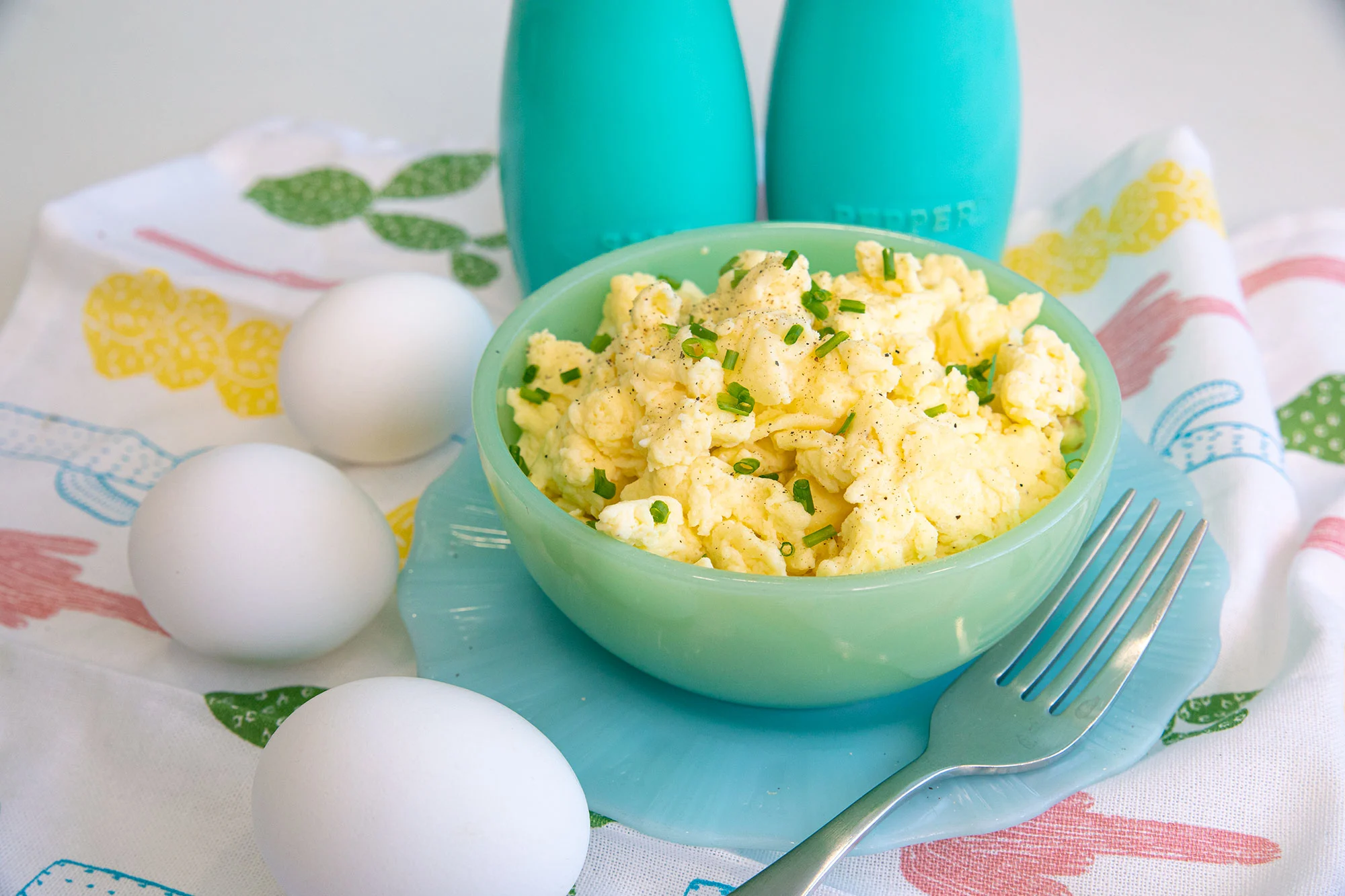 A bowl of scrambled eggs with chives, three whole eggs, a fork, and blue salt and pepper shakers.