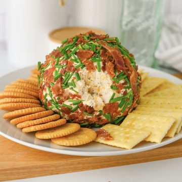 A ranch cheese ball coated with bacon and chives, served with round and square crackers on a white plate.