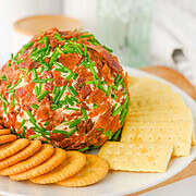 A ranch-flavored cheese ball coated in bacon and chives, served with round and square crackers on a white plate.