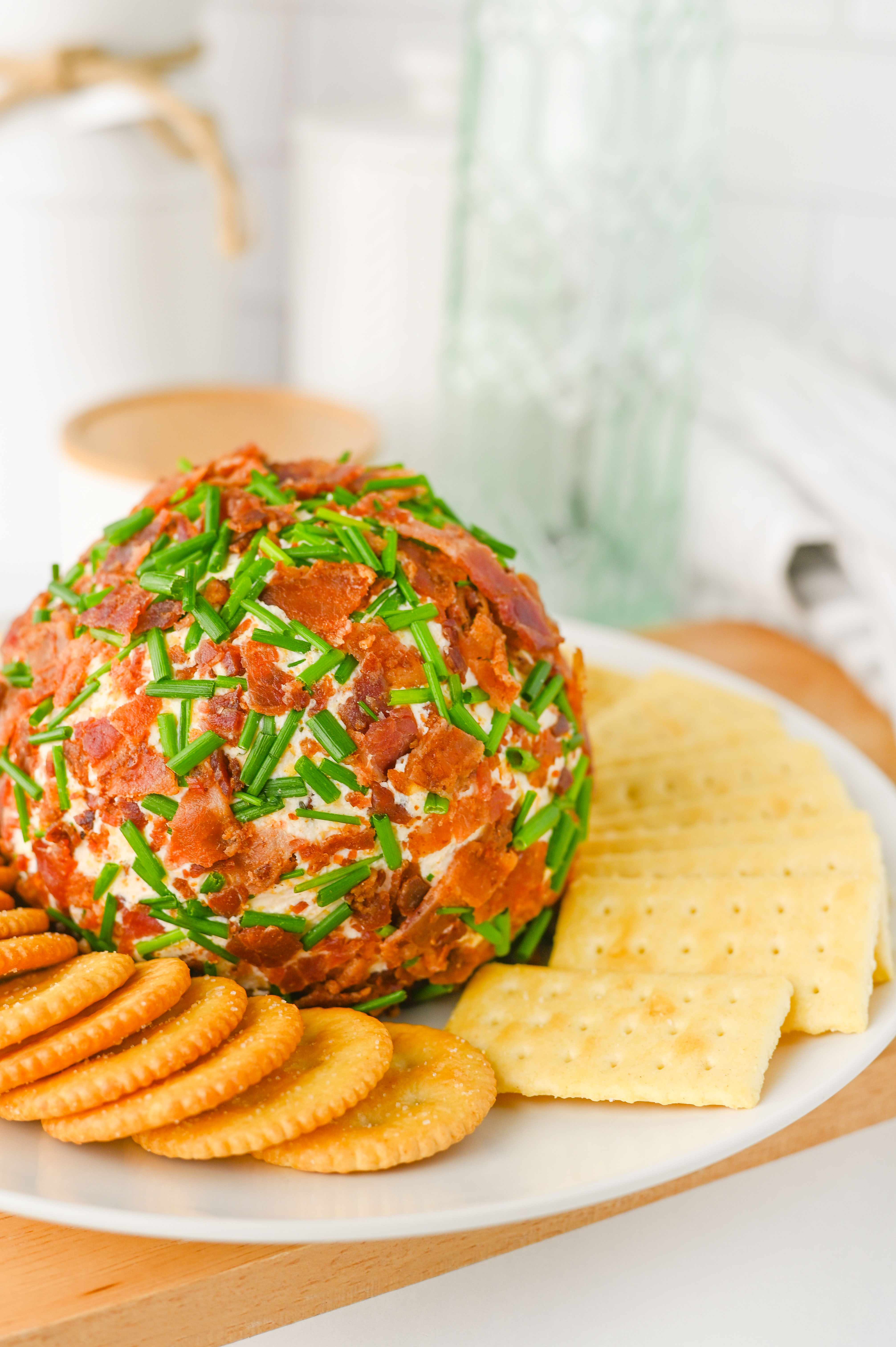 A ranch-flavored cheese ball coated in bacon and chives, served with round and square crackers on a white plate.