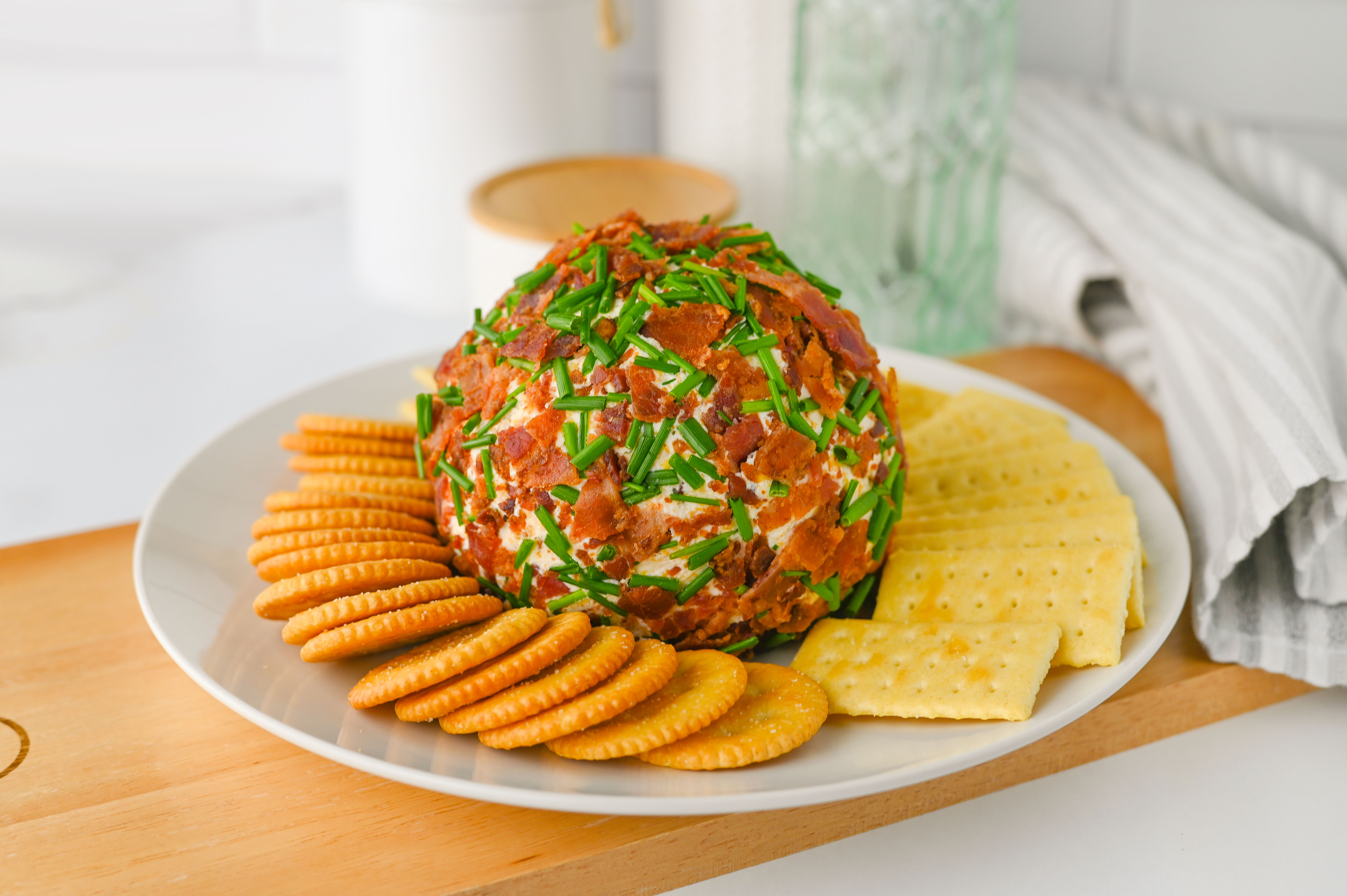 A ranch-infused cheese ball coated with crispy bacon and fresh chives, served with round and square crackers on a white plate.