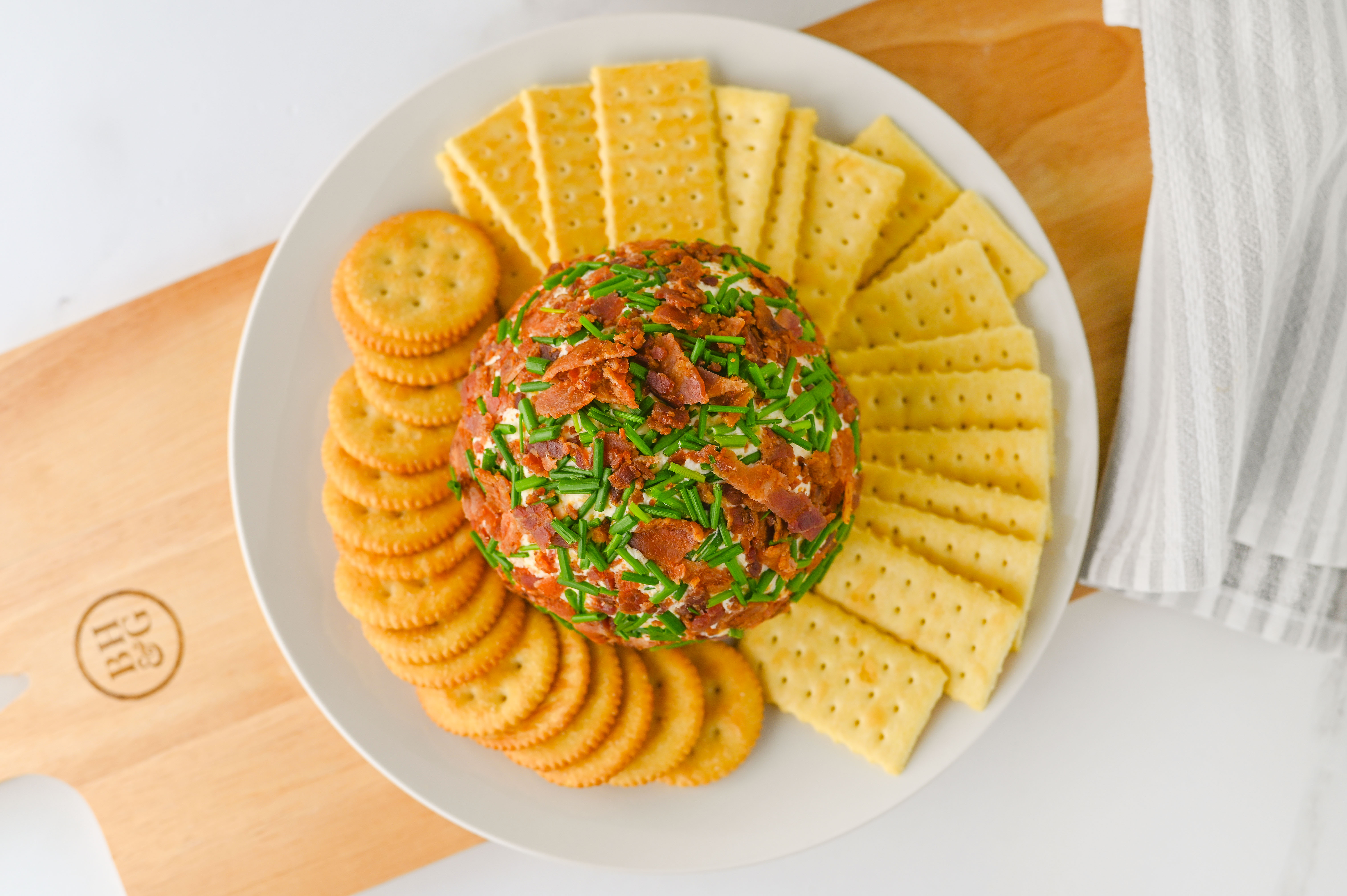 A ranch-infused cheese ball coated with bacon and chives, served with round and rectangular crackers on a white plate.