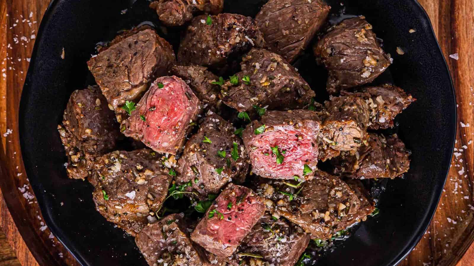 Steak bites topped with herbs and coarse salt on a black plate, some pieces are medium-rare.