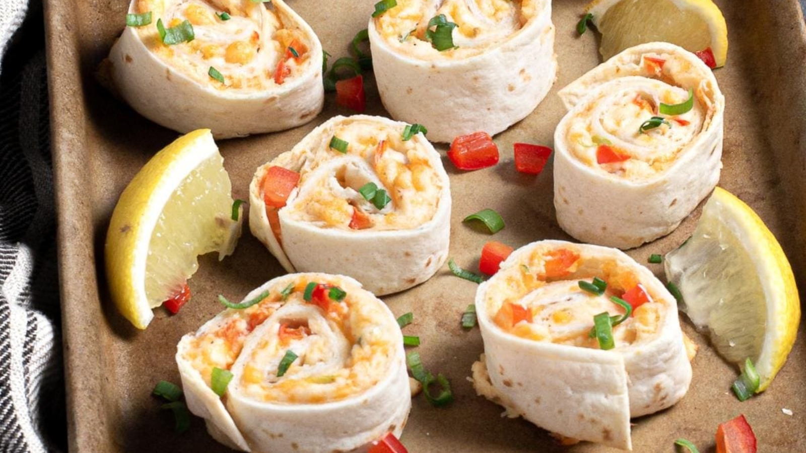 Six tortilla pinwheels with cheese and peppers, garnished with green onions and lemon wedges on a tray.
