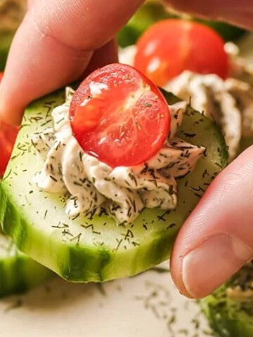 A hand holding a cucumber slice topped with herbed cream cheese and a cherry tomato half.
