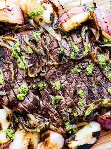Grilled steak topped with green sauce, surrounded by grilled onions and potatoes on a wooden board.