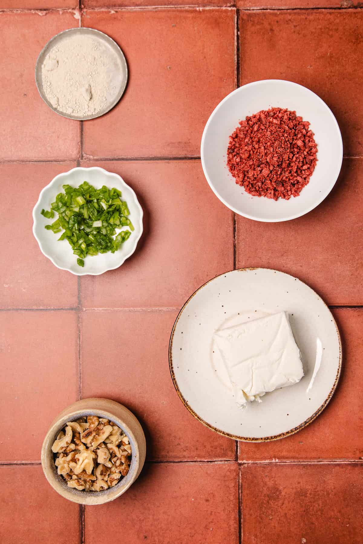 Five bowls display ingredients for a southern cream cheese ball recipe: chopped walnuts, green peppers, white powder, red bits, and cream cheese, all arranged on a vibrant red tiled surface.