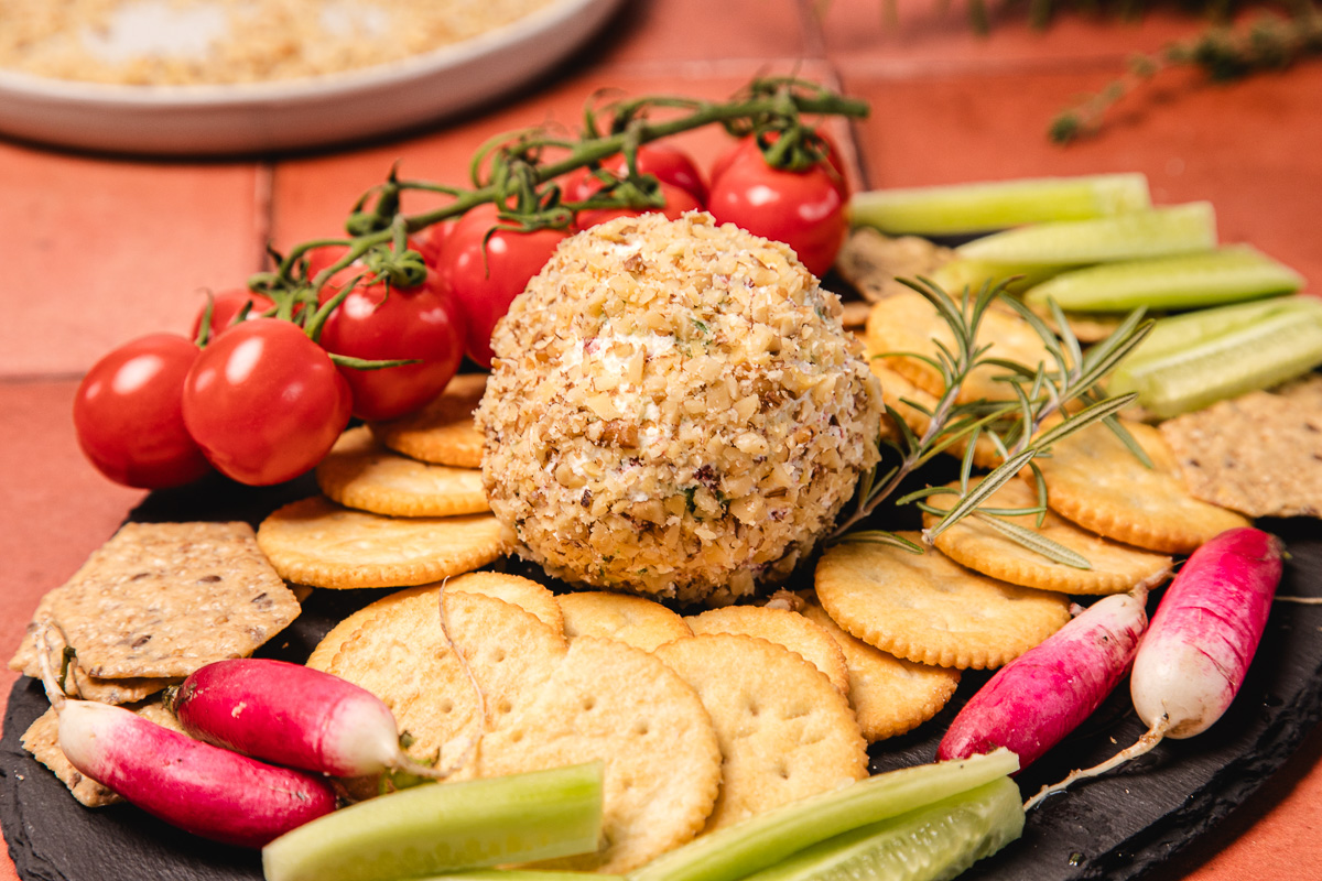 A southern cream cheese ball recipe coated in nuts sits on a platter, surrounded by crackers, cherry tomatoes, cucumber, and radishes.