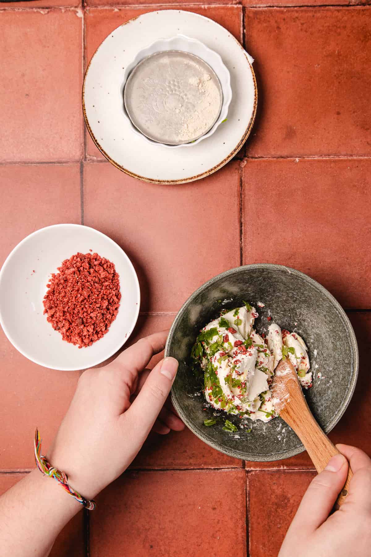 A person mixes herbs into cream cheese in a bowl, preparing a southern cream cheese ball recipe, with bowls of dried herbs and pink seasoning nearby.