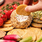 A hand dips a cracker into a southern cream cheese ball recipe, surrounded by crackers, tomatoes, radishes, and celery.