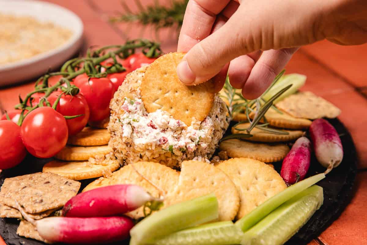 A hand dips a round cracker into a southern cream cheese ball recipe, surrounded by crackers, tomatoes, radishes, and cucumber.
