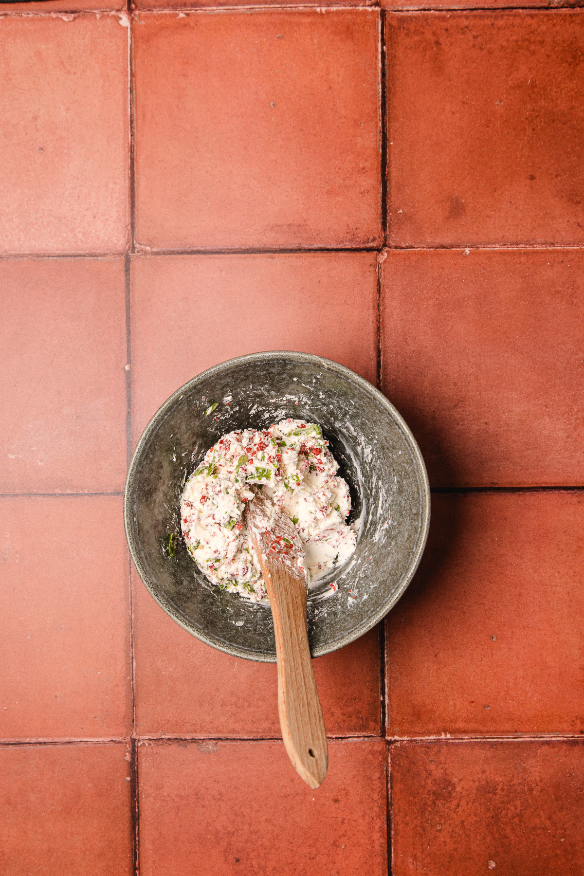 A bowl of herbed dough with a wooden spoon on a red tiled surface, seen from above—perfect for creating your favorite southern cream cheese ball recipe.