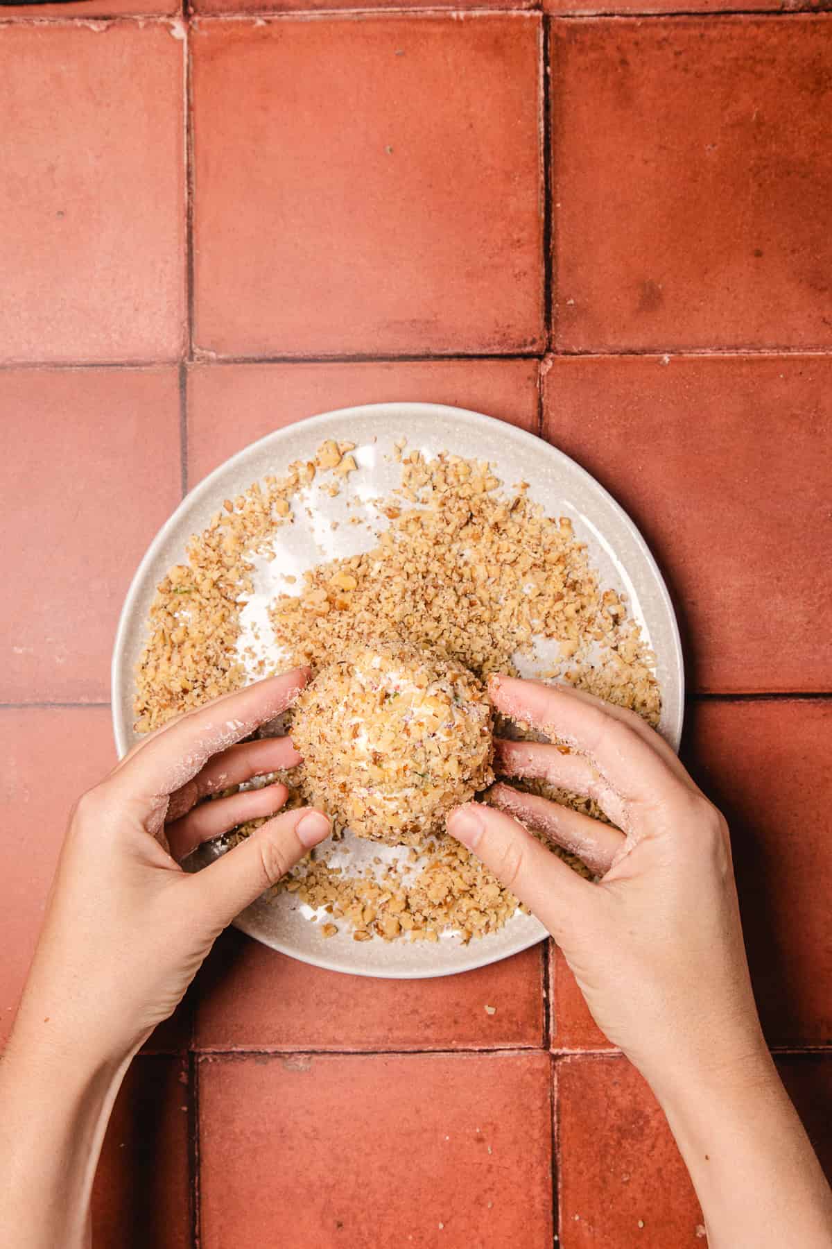 Hands rolling a round snack in crushed nuts on a white plate, placed on a red tiled surface, perfect for your next southern cream cheese ball recipe.