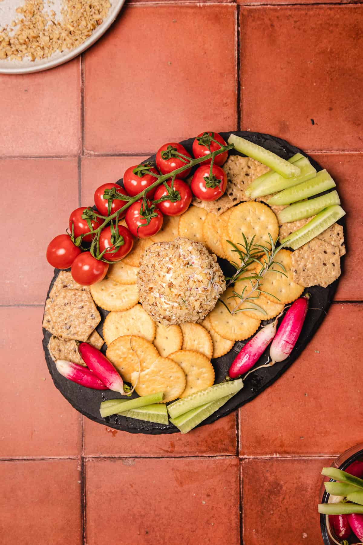A southern cream cheese ball recipe served with crackers, cherry tomatoes, celery, and radishes on a black platter over a red tiled surface.