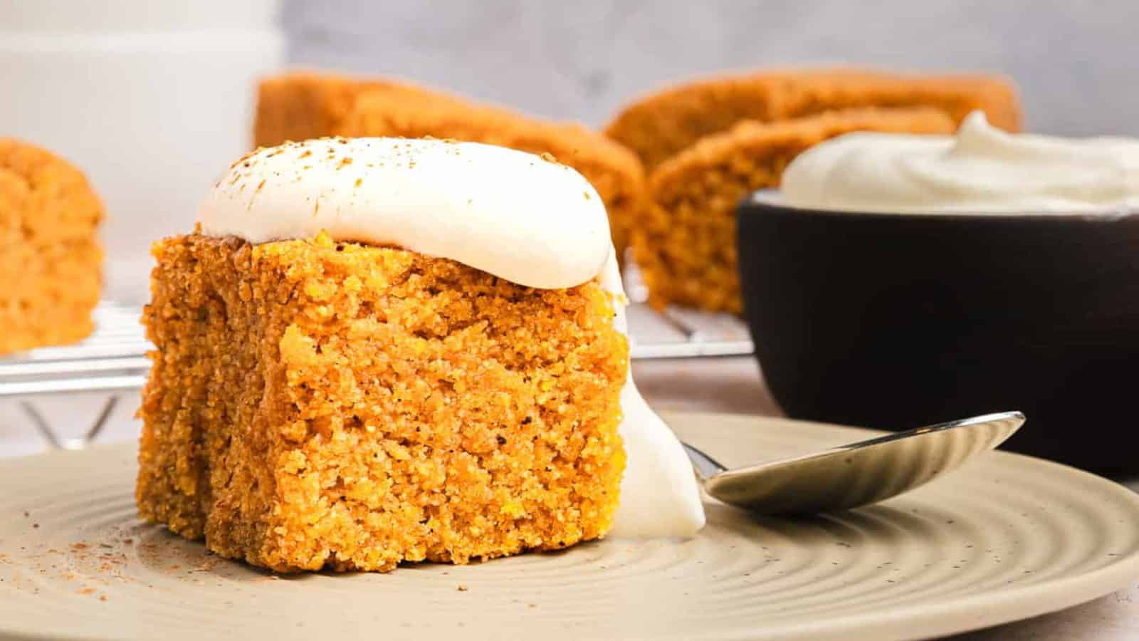 A slice of spiced cake with creamy frosting on a plate, with more cake and a bowl of frosting in the background.