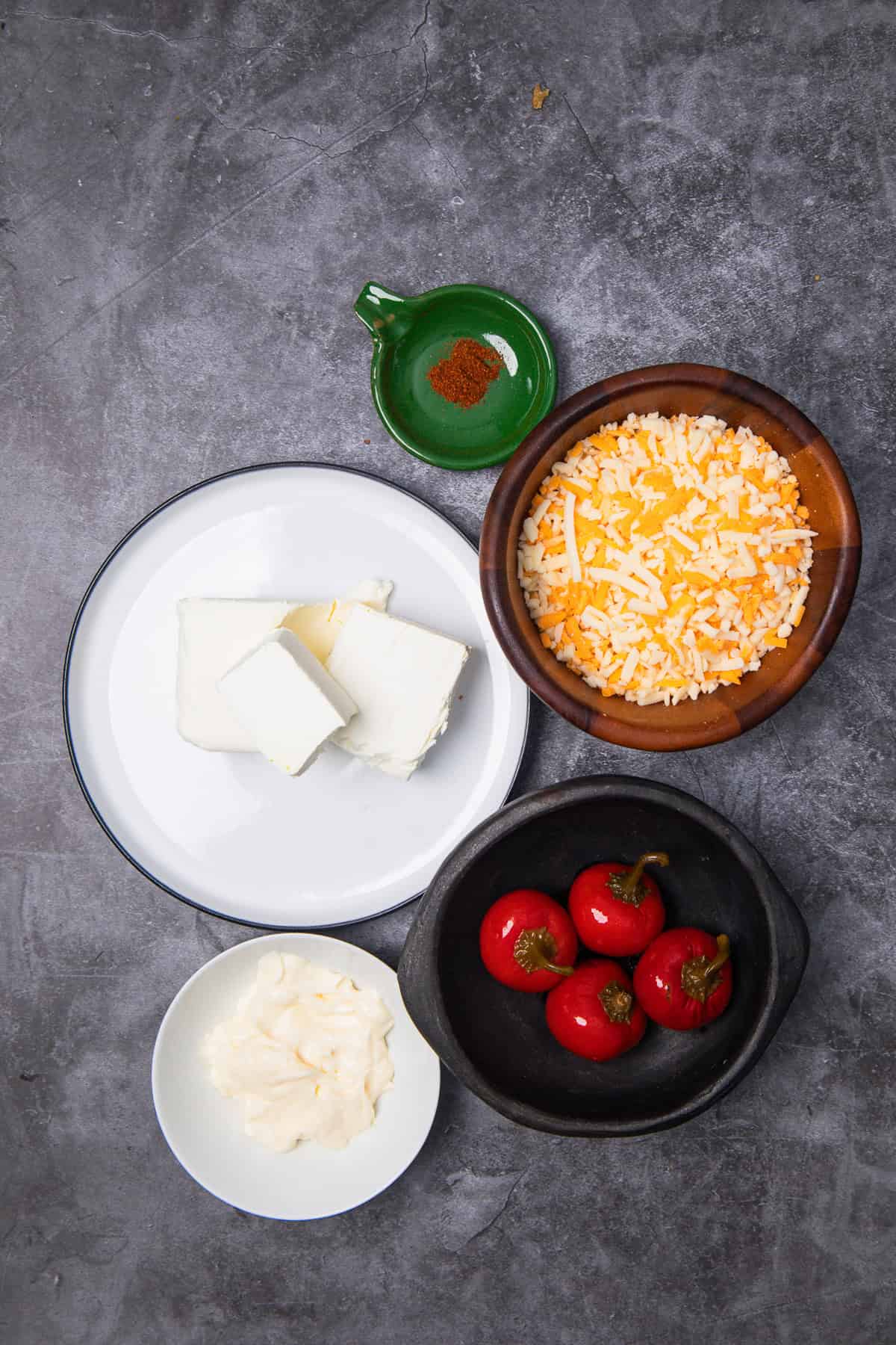 Four bowls of cream cheese, shredded cheese, mayo, red peppers, and a small dish of spices on a gray surface—everything you need for a classic Southern Pimiento Cheese dip.