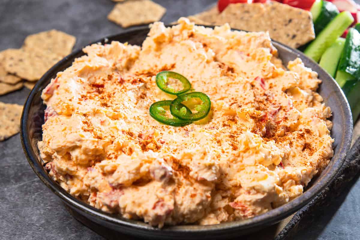 A bowl of vintage Southern cheese dip topped with sliced jalapeños, surrounded by crackers and fresh vegetables.