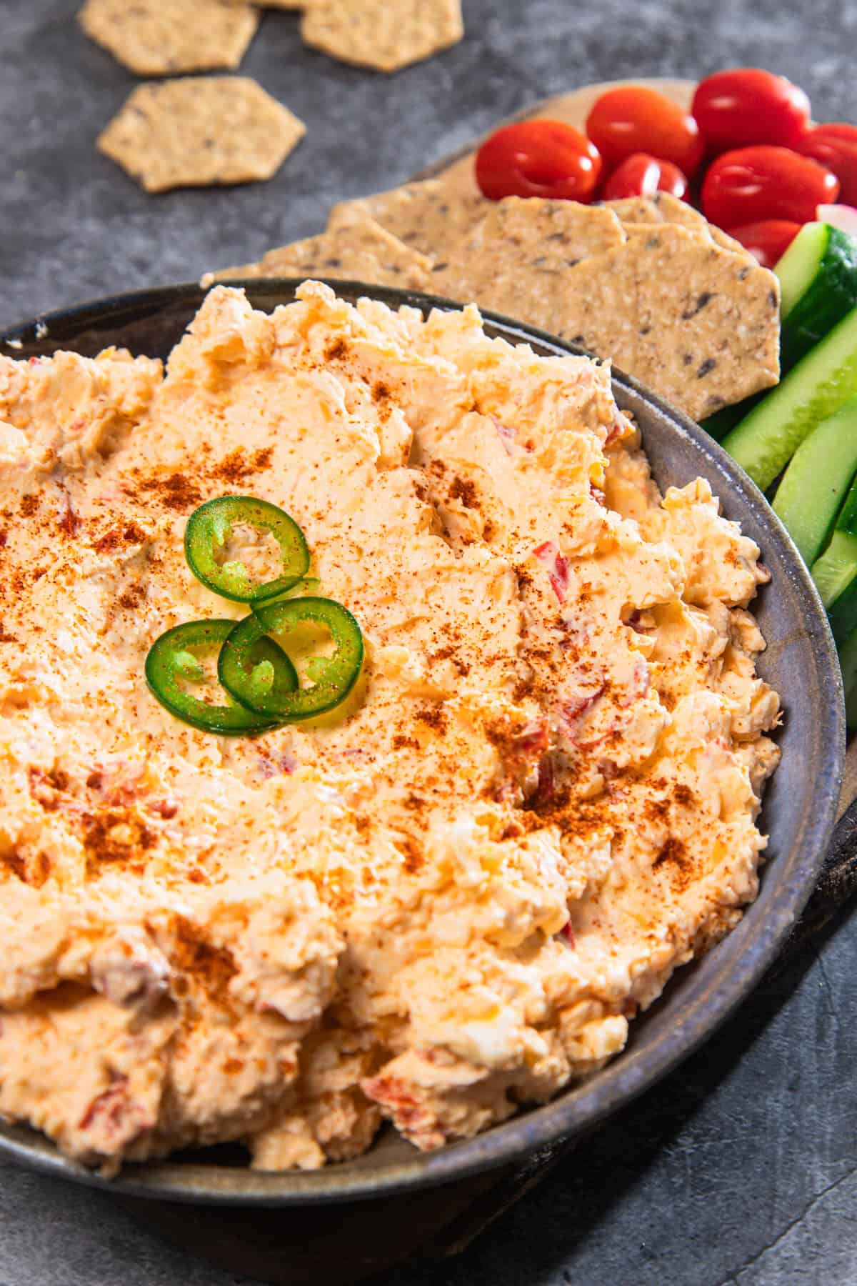 A bowl of vintage Southern pimiento cheese dip topped with jalapeño slices, served with crackers, tomatoes, and cucumber sticks.