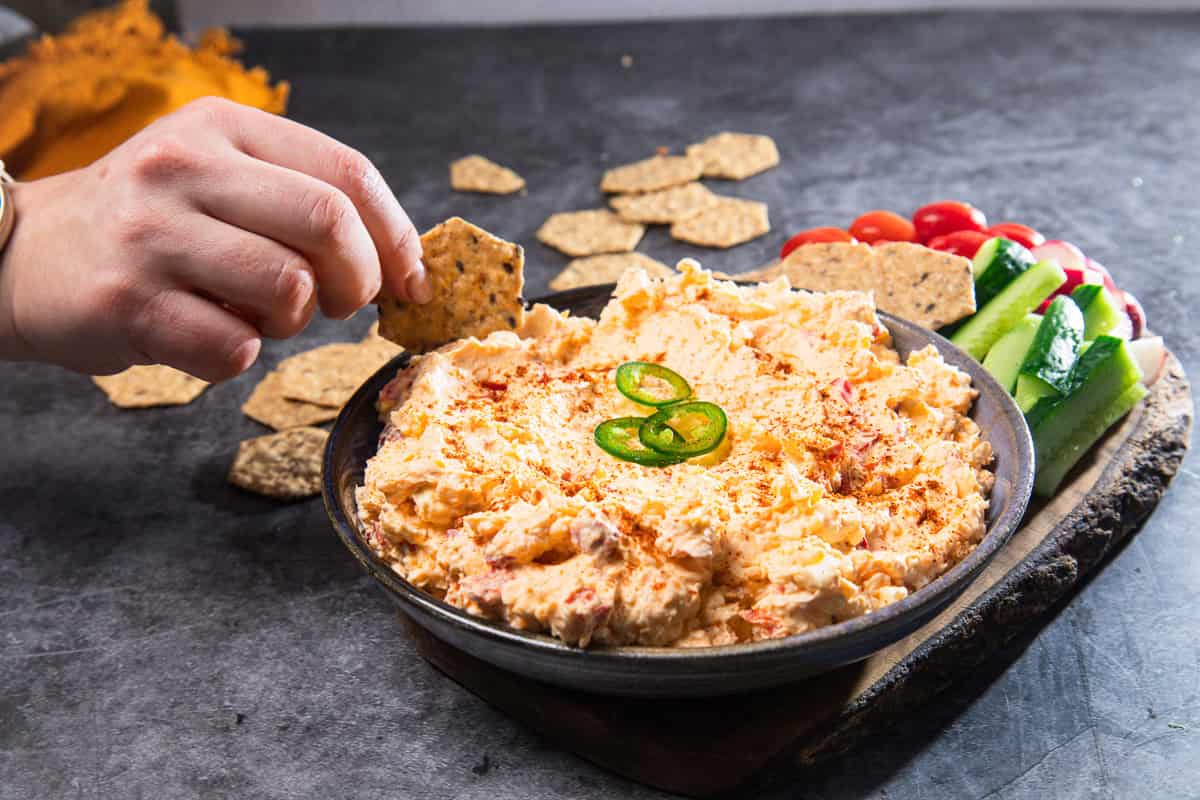 A hand dips a cracker into a bowl of Southern Pimiento Cheese dip, with veggies and more crackers on the side.