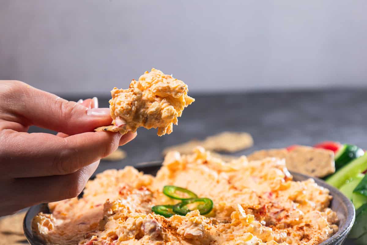 A hand holds a chip with creamy Southern Cheese Dip above a bowl of dip topped with sliced jalapeños.