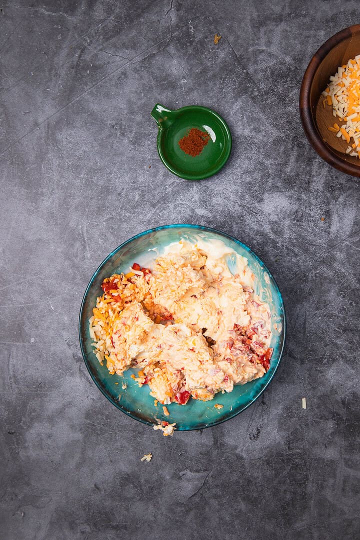 A bowl of shredded cheese and creamy Cheese Dip mixture rests on a gray surface, evoking Vintage Southern charm, with a small green dish of paprika nearby.