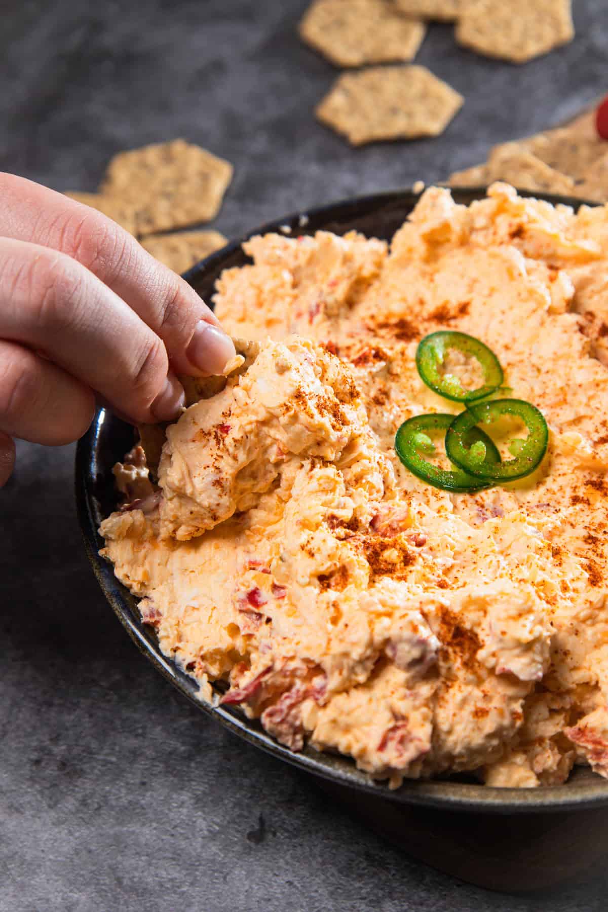 A hand dips a cracker into a bowl of creamy Southern Pimiento Cheese Dip, topped with sliced jalapeños and paprika for a vintage-inspired snack.