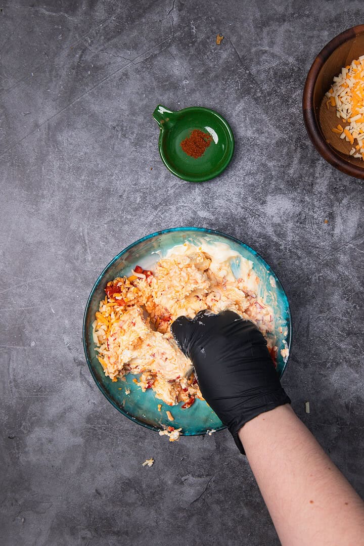 A gloved hand mixing shredded cheese and ingredients in a bowl on a gray surface, crafting a classic Vintage Southern pimiento cheese dip, with a small dish of paprika nearby.