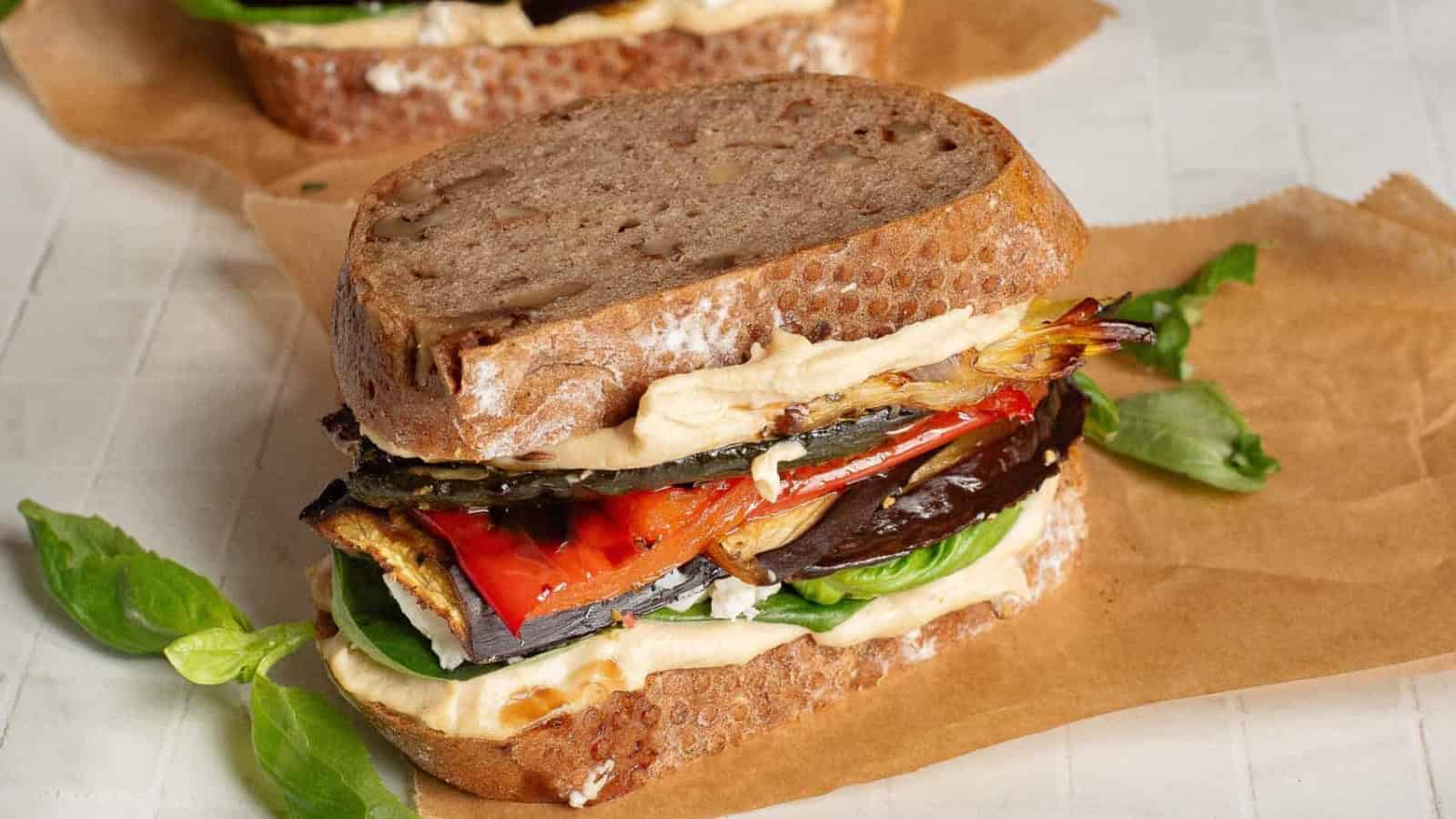 A sandwich with hummus, roasted vegetables, and lettuce on rustic bread, placed on parchment paper.