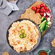 Bowl of Vintage Southern Pimiento Cheese Dip topped with jalapeƱos, served with crackers, cherry tomatoes, cucumbers, and radishes.