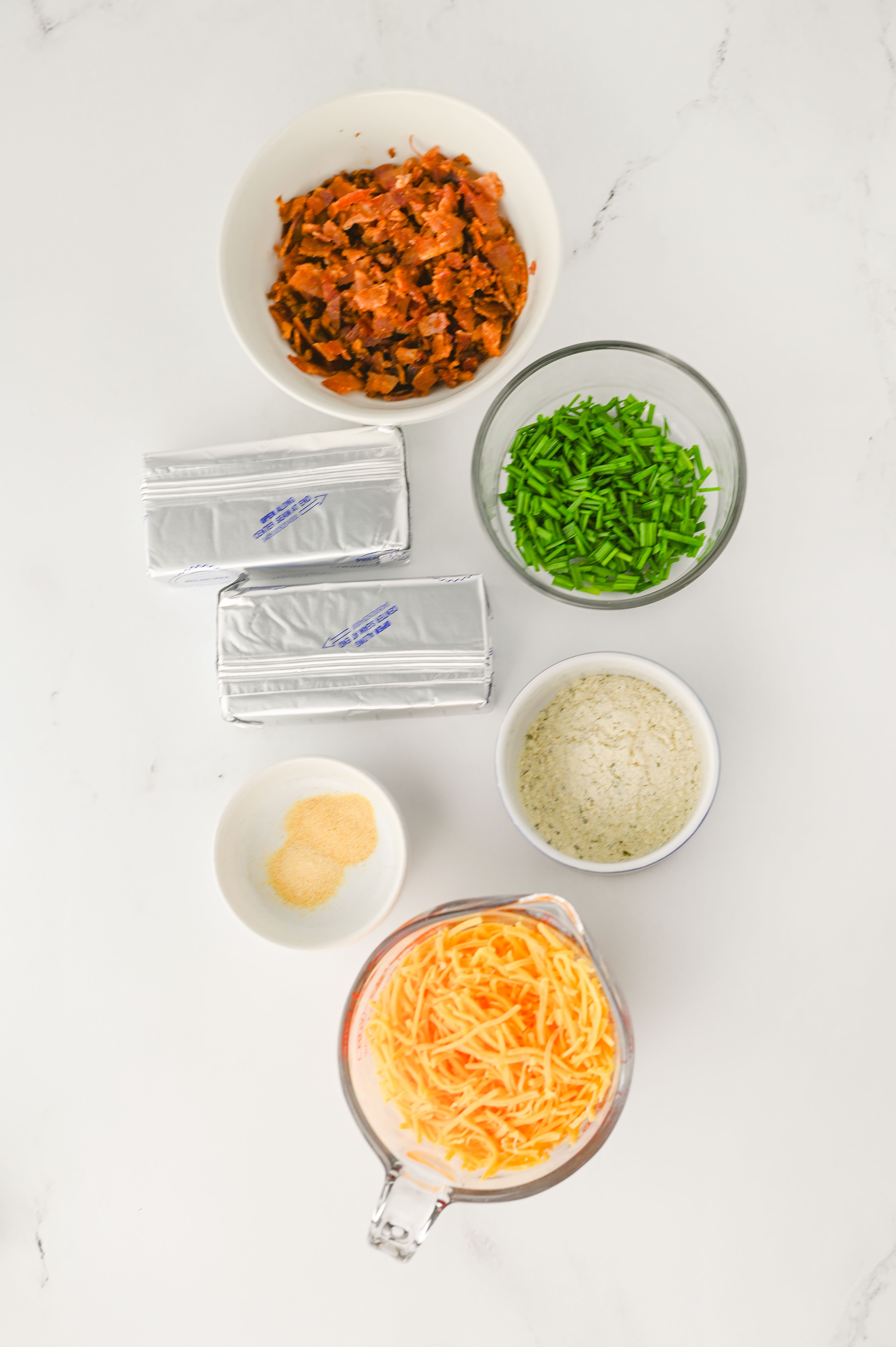 Ingredients for Bacon Ranch Cheese Balls, including bacon bits, chives, cream cheese, seasonings, and shredded cheese, are arranged on a white surface.