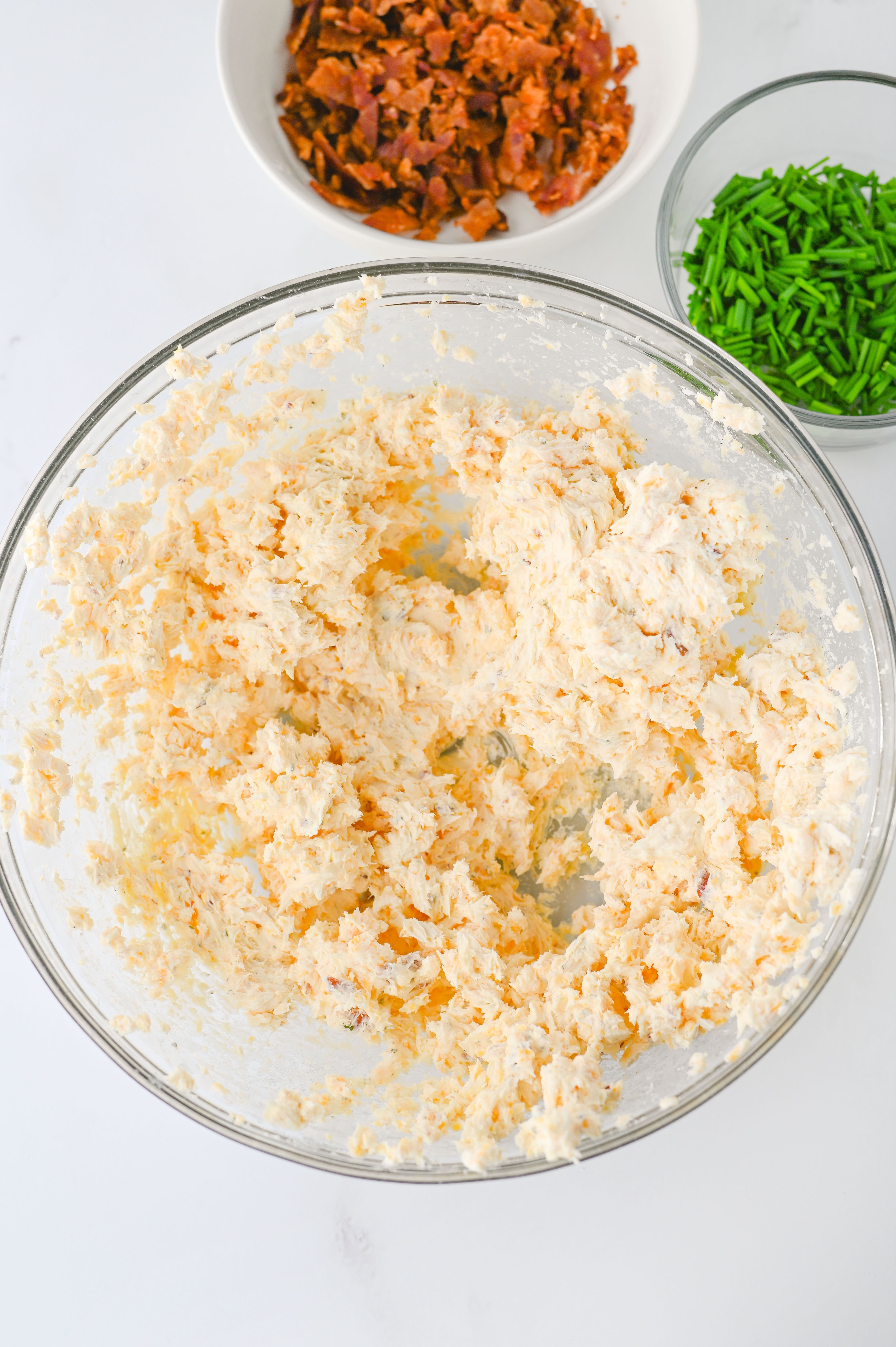 A glass bowl of creamy ranch cheese mixture, with bowls of chopped bacon and chives in the background—perfect for making delicious cheese balls.