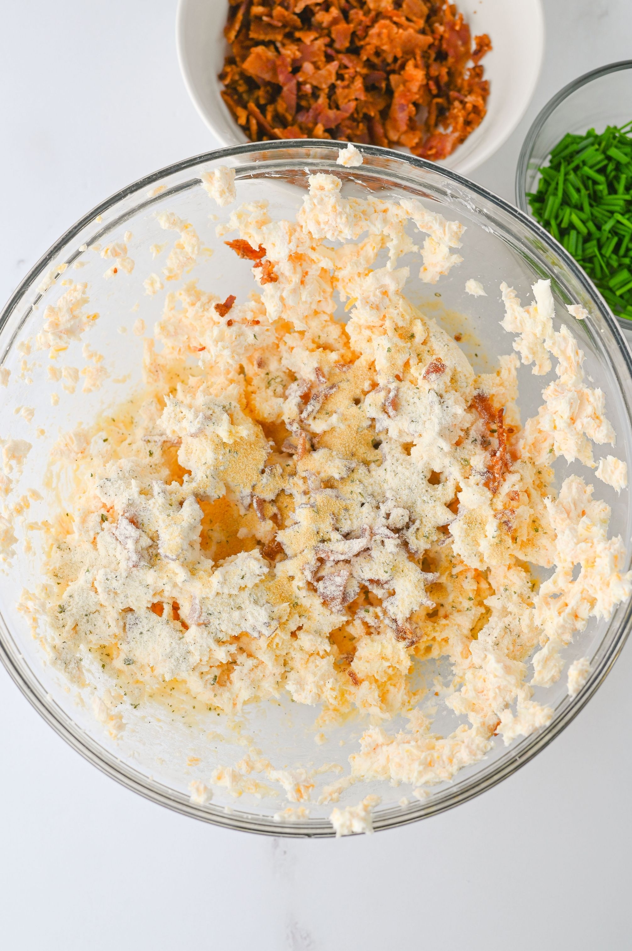 Creamy mixture in a glass bowl with crumbled bacon; chopped chives in a bowl nearby, perfect for making flavorful bacon ranch cheese balls.