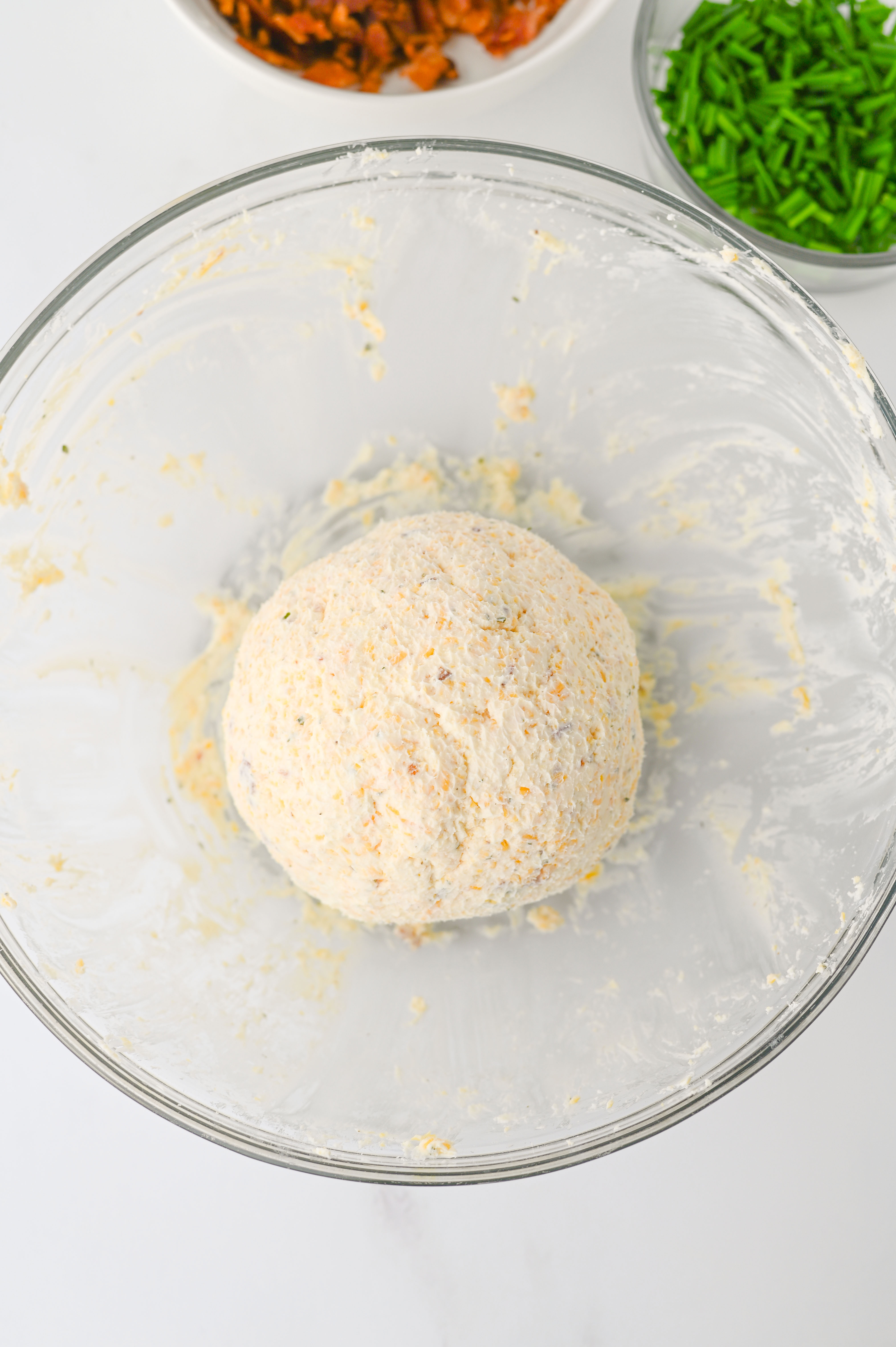 A ball of dough in a glass bowl, with bowls of chopped chives, bacon, and other ingredients nearby—perfect for crafting savory ranch cheese balls.