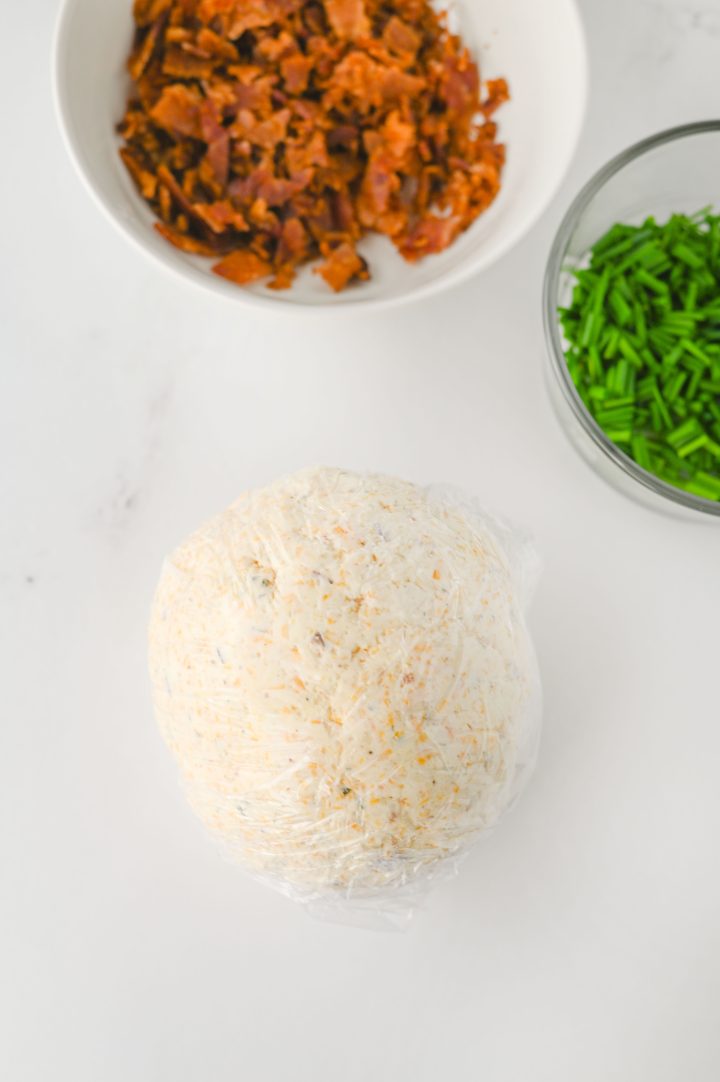 Cheese ball wrapped in plastic wrap, with bowls of chopped bacon and chives nearby on a white surface, perfect for making flavorful ranch cheese balls.