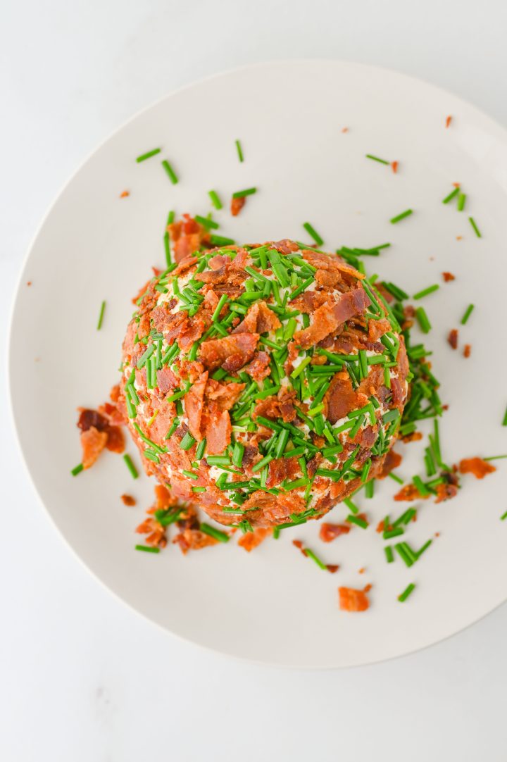 A ranch-flavored cheese ball covered with chopped bacon and chives sits on a white plate.
