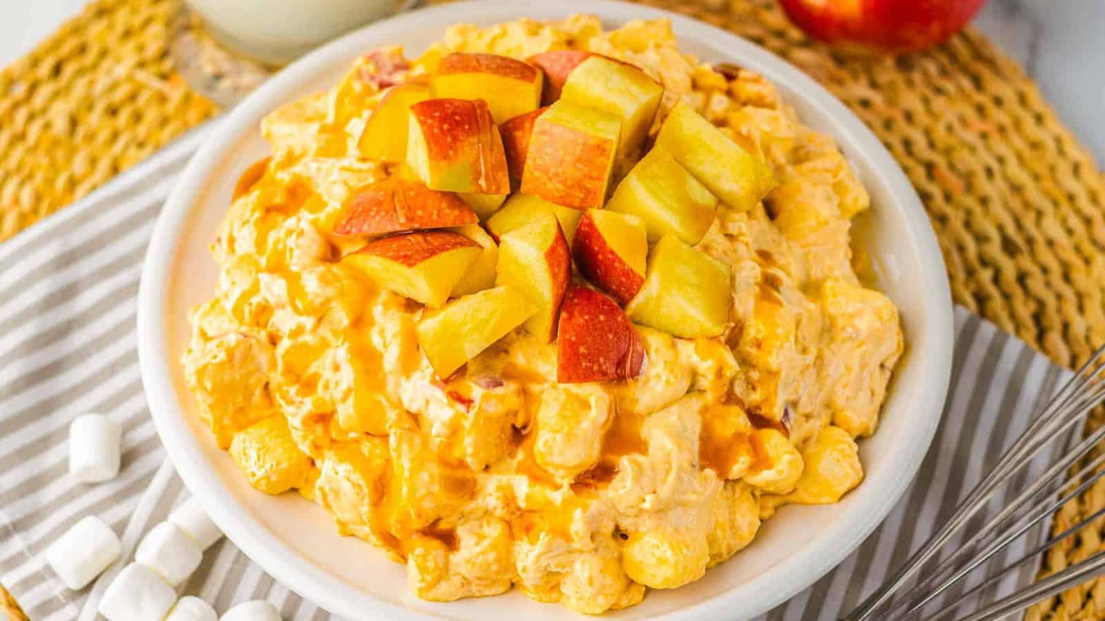 A bowl of creamy salad topped with chopped apples, surrounded by marshmallows on a striped napkin.