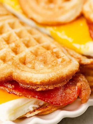 Waffle sandwich with egg, cheddar cheese, and crispy bacon on a white plate.