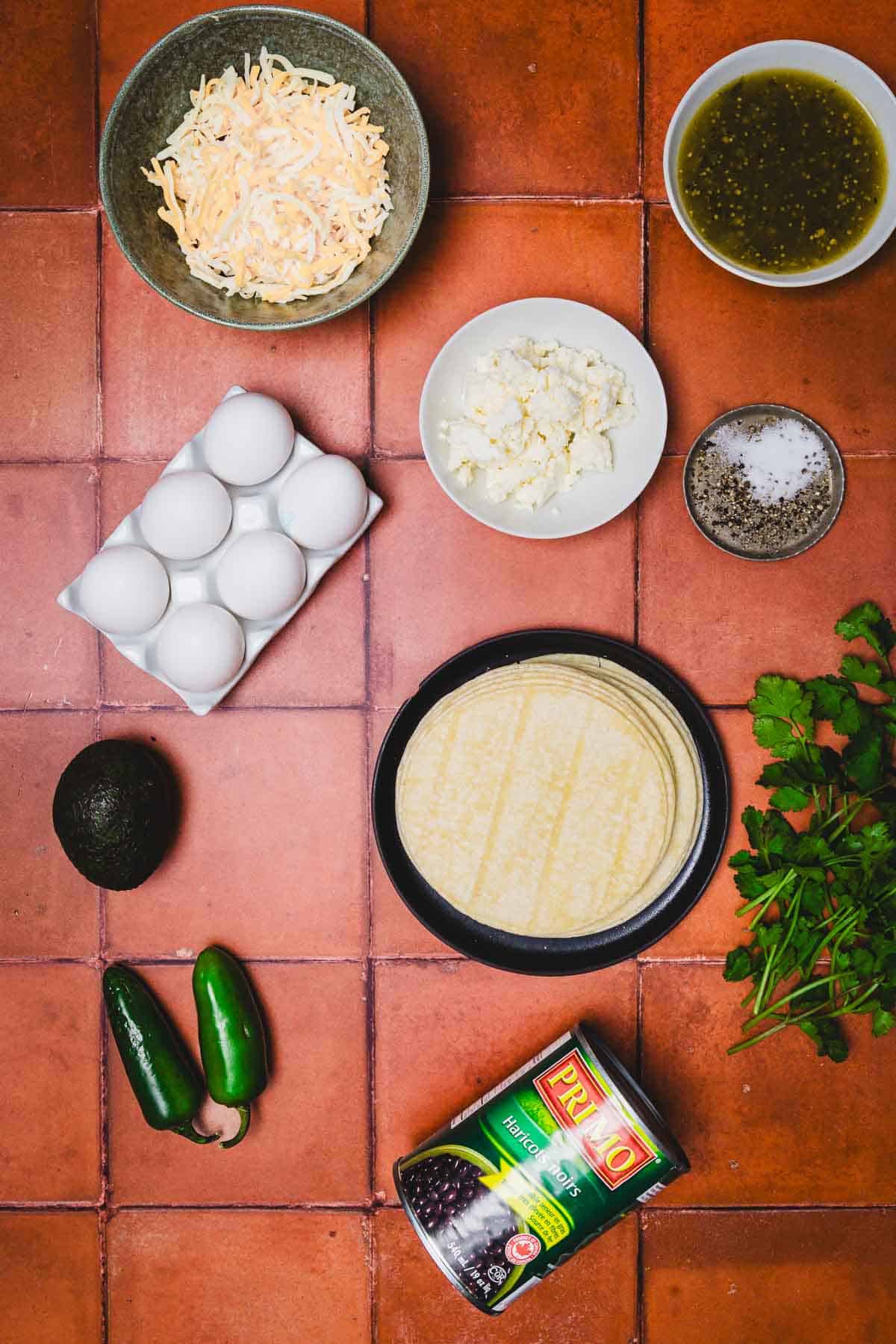 Tortillas, eggs, cheese, salsa, cilantro, avocado, jalape&ntilde;os, black beans, and salt come together on a tile surface for a vibrant huevos rancheros breakfast casserole.