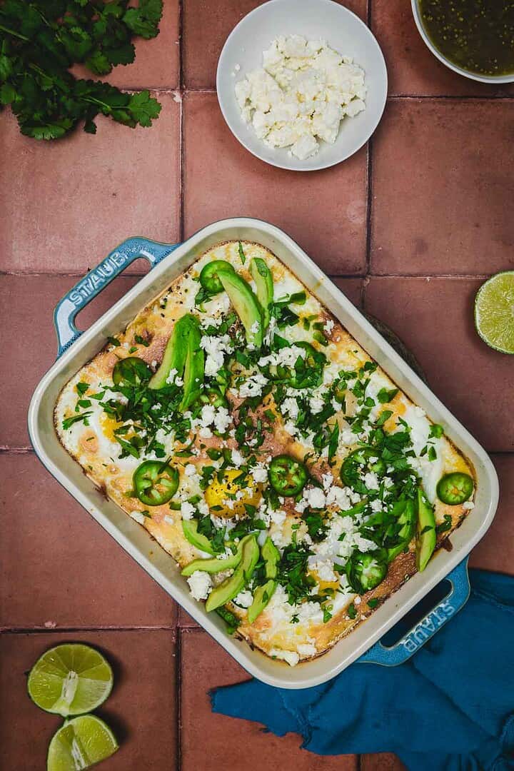 Huevos rancheros breakfast casserole baked to perfection and topped with avocado, jalape&ntilde;os, cheese, and fresh cilantro; served with lime and feta on the side, a blue towel nearby.
