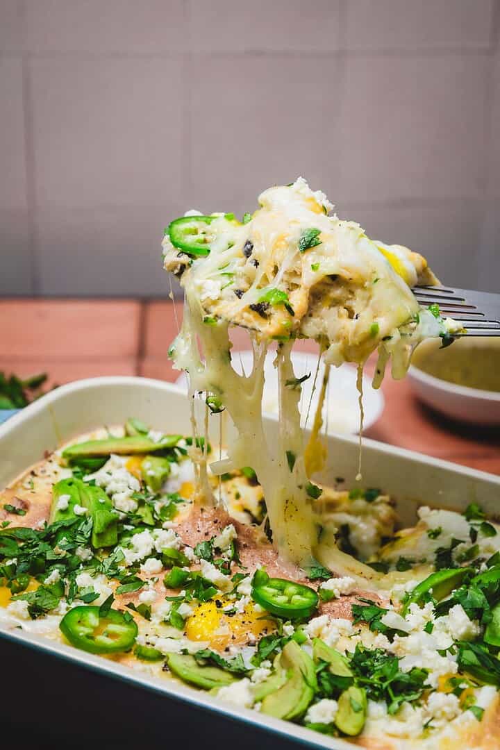 A fork lifts a cheesy, baked huevos rancheros breakfast casserole topped with jalape&ntilde;os, herbs, and crumbled cheese from a dish.